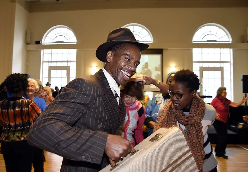 A man dressed in an old-fashioned pin stripe suit and trilby, smiles and dances with a suitcase