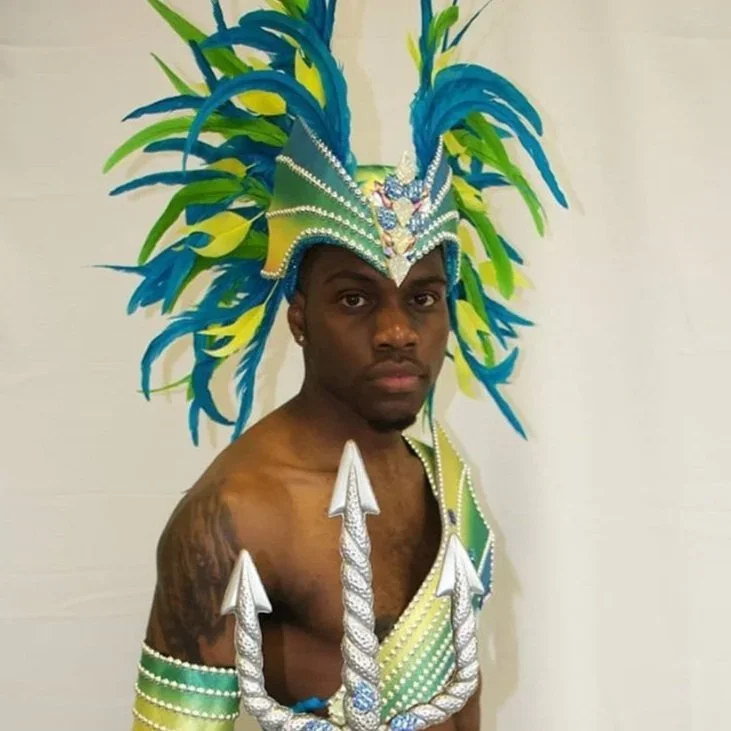 A man in a green and blue dragon-style headdress holds a silver trident and looks into the camera
