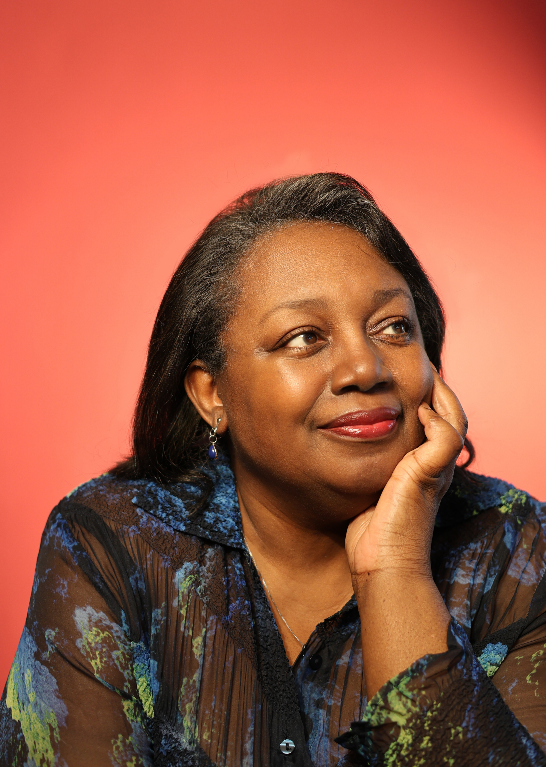 A black woman smiling and posing with her hand on her face, against a bright orange and pink ombre backdrop.