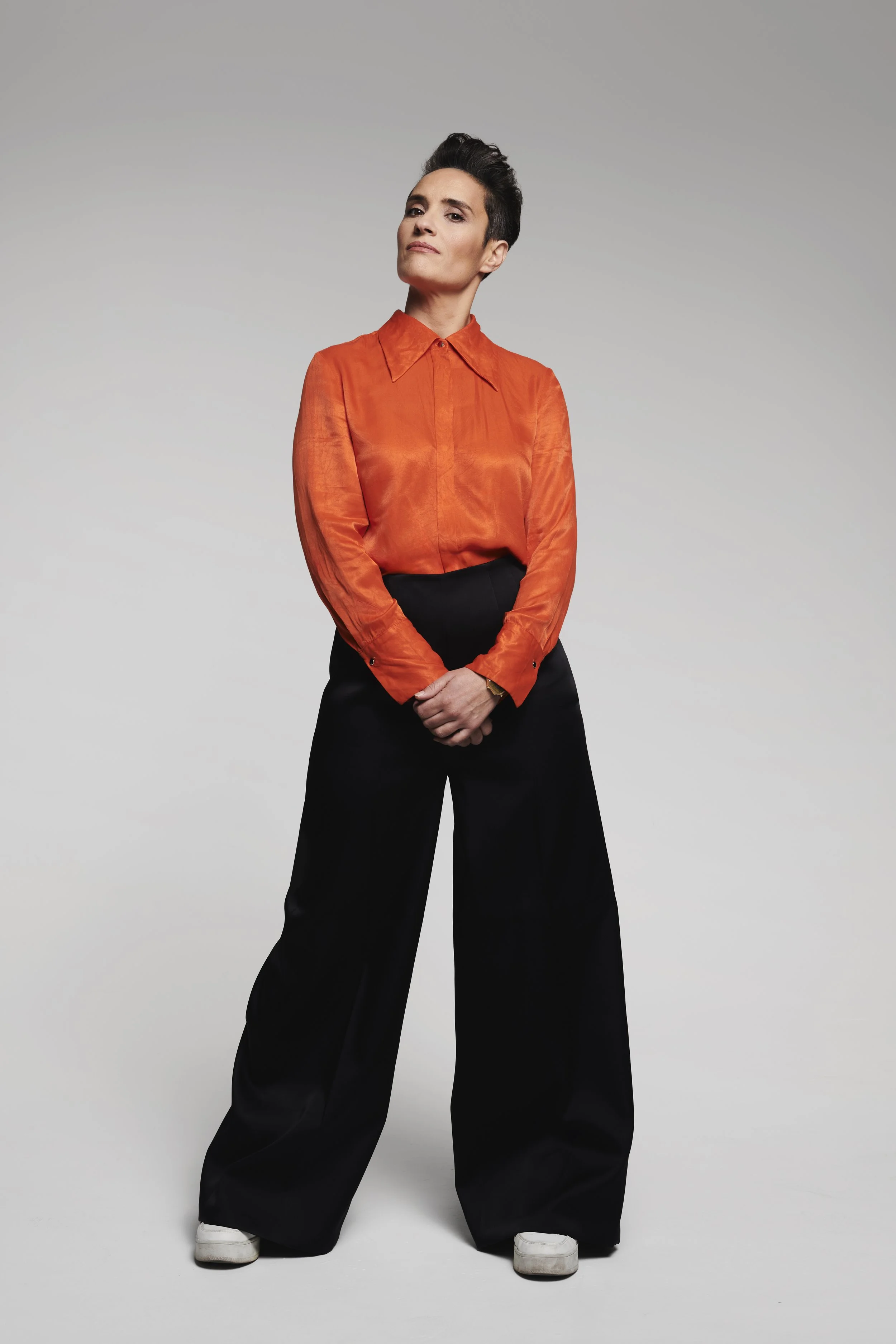 A photo of Jen Brister, a white woman standing in an orange shirt, black trousers and white trainers.