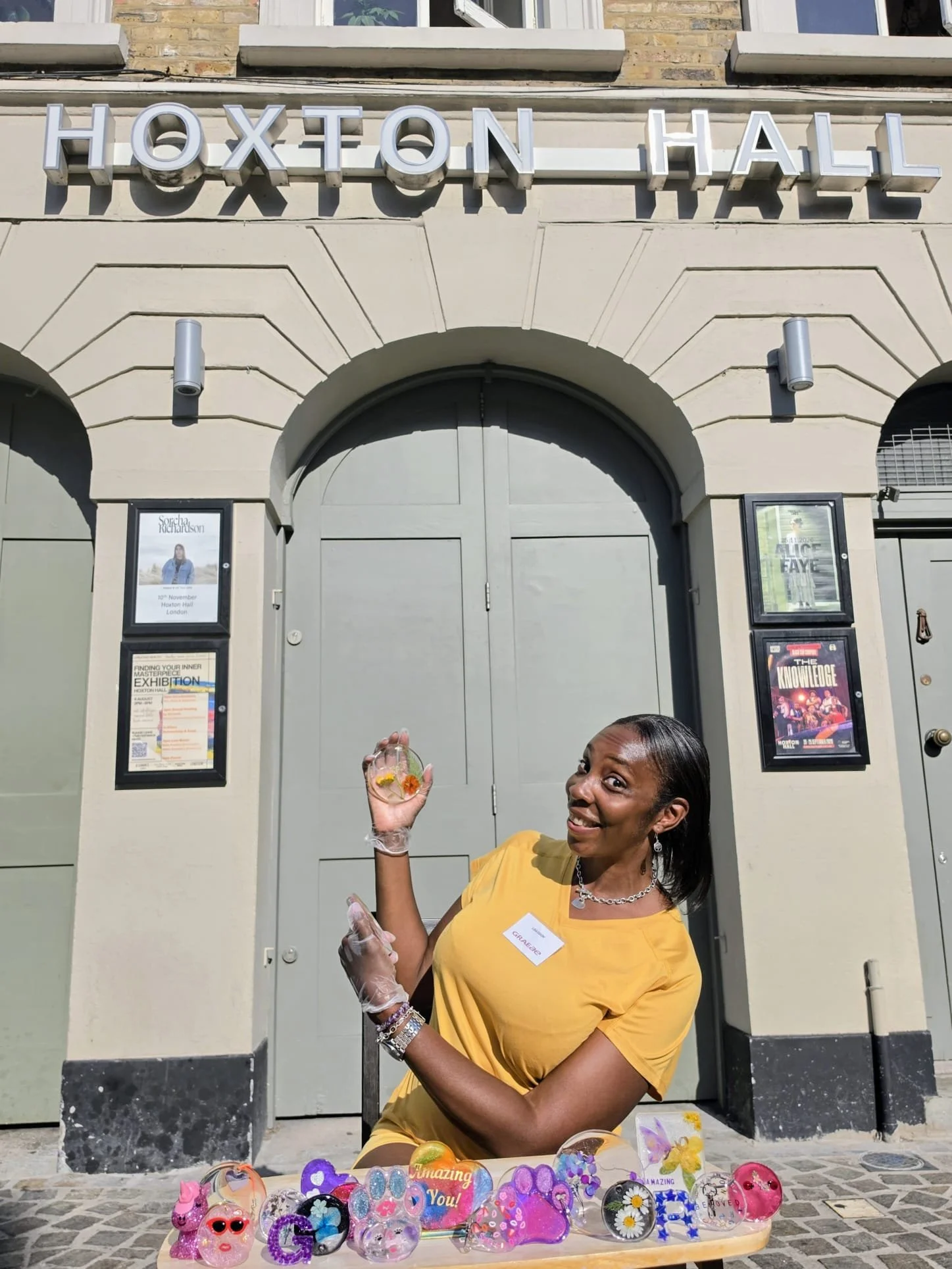 A black woman in a bright yellow t-shirt sits infant of Hoxton Hall with a ra age of resin items on a table