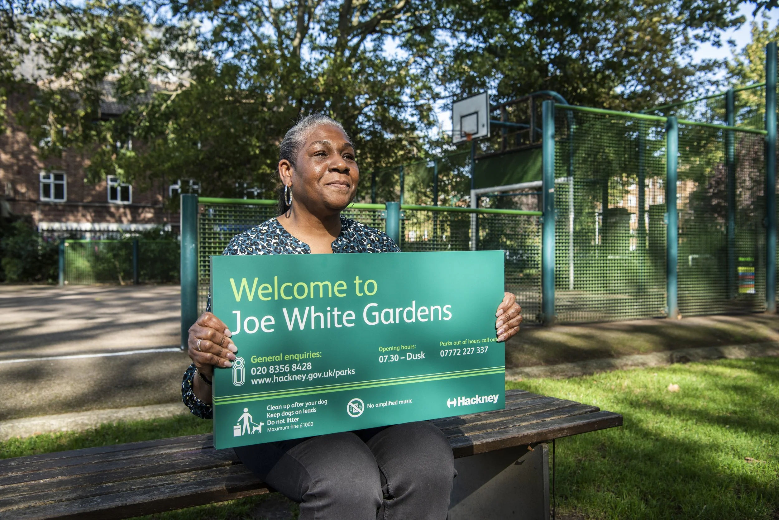 Black Woman holding a Hackney park's sign that reads 'welcome to Joe White Gardens'