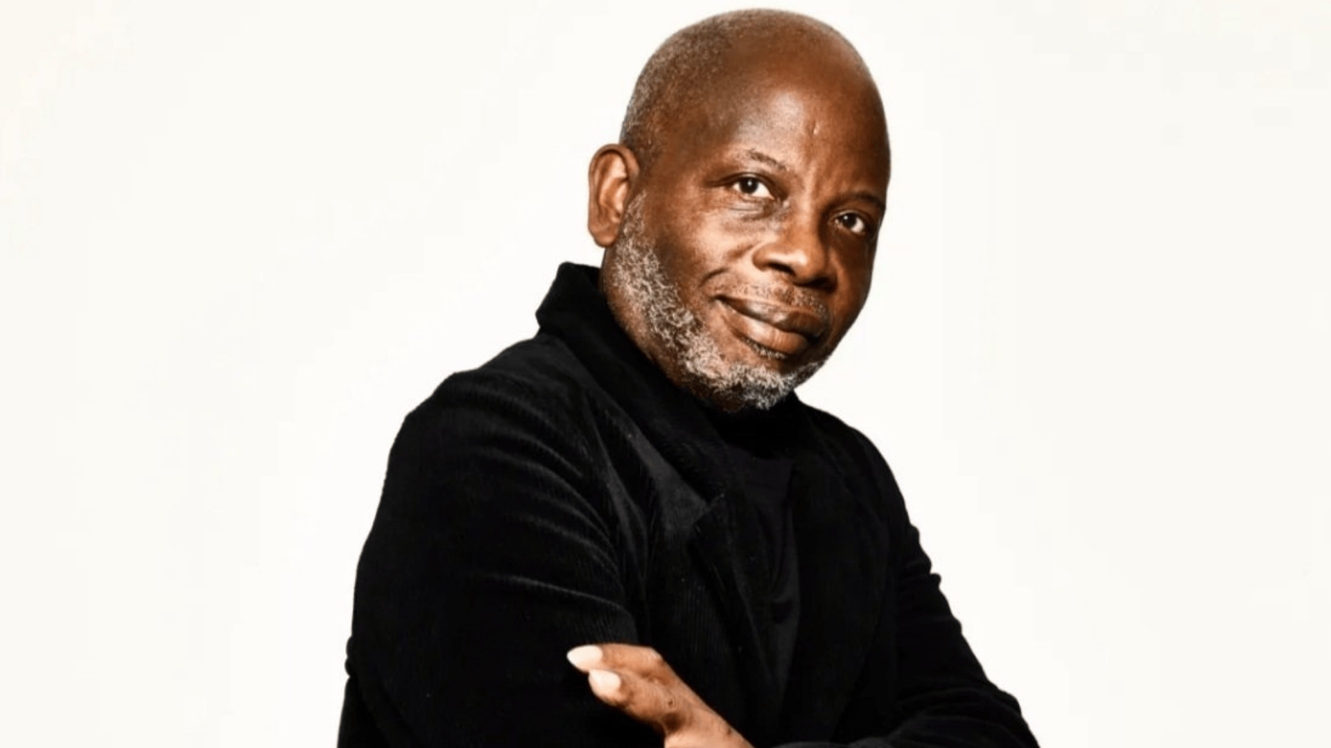 A Black man with a beard wearing a black top smiles at the camera against a white background