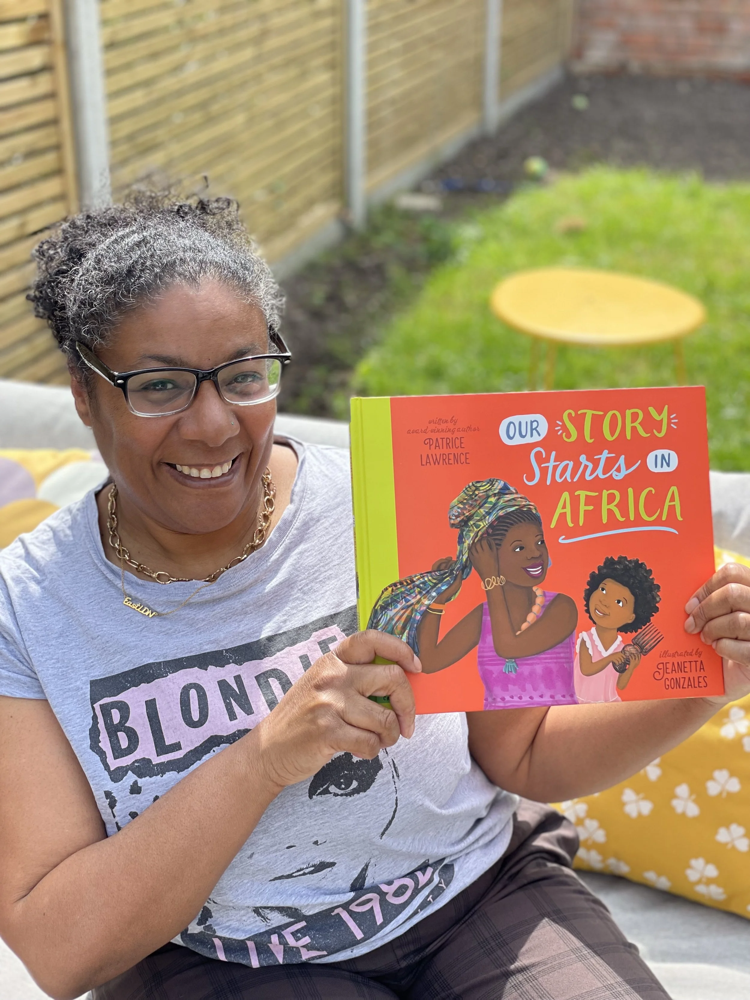 Author Patrice Lawrence smiling and holding her bright red book, Our Story Starts in Africa, against a garden backdrop.