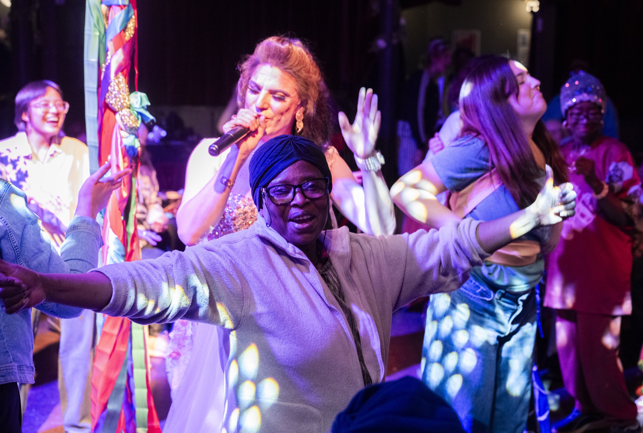An older woman dances with her arms outstretched in a joyous moment, while a drag artist sings into a mic behind her.