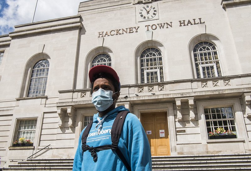 A man wearing a face mask, blue hoodie and red cap stands in front of Hackney Town Hall.