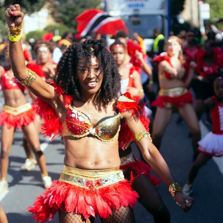 A woman walks in a parade, she's wearing a gold and red bra and skirt with red feathers and hold her right hand aloft. She has long black afro-style hair.