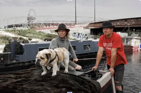 Two people and a dog stand by a lock as a canal boat sits in the background.