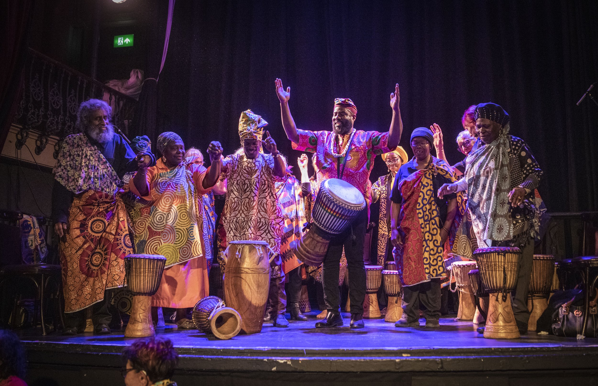 A community band of djembe drummers stand on a stage in African costume.  The band leader lifts his hands in celebration.