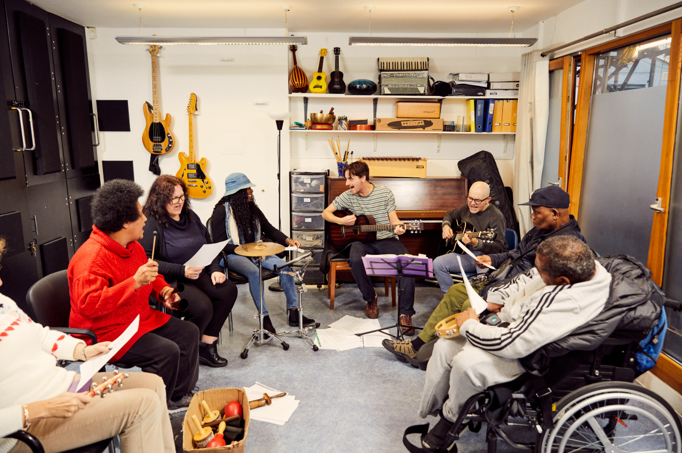 A group of people of all abilities sit in a semi-circle in a music room and play music together.