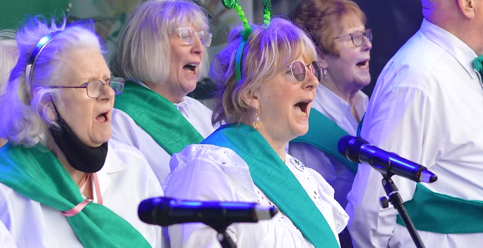 a group of irish elders sing in a choir, wearing white shirts and green sashes
