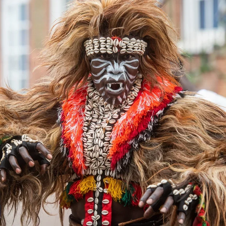 A man dressed an African-style lion costume with a long fur coat and shell headdress and breastplate. His face is painted with white lines as if depicting whiskers and sharp teeth
