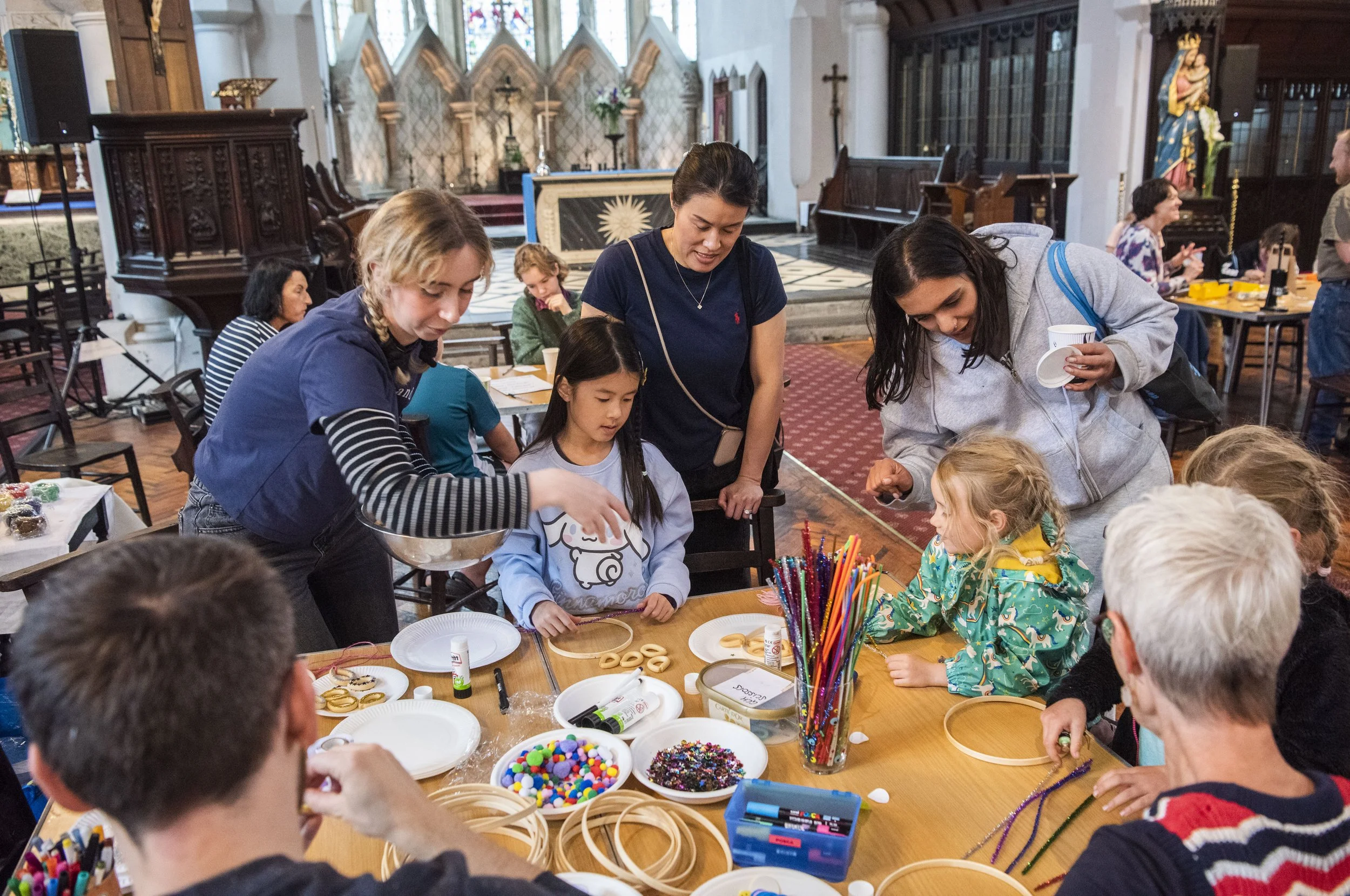 A group of white peole of al ages sit around a table in a church, involved in a creative workshop