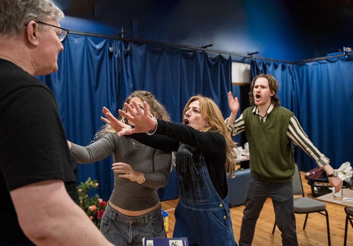 4 people are in a rehearsal room with blue curtained walls, one man with grey hair has his back to us, 2 people stand close with their hands outstretched facing the camera with another person shouting behind them