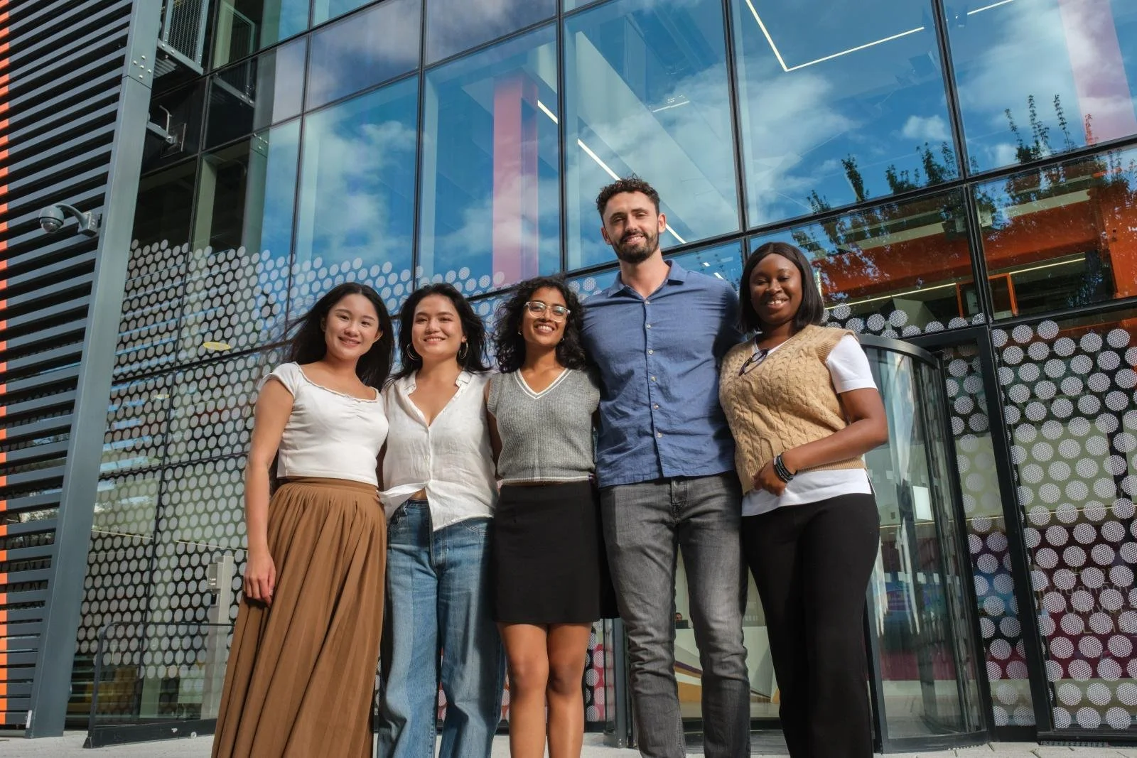 Five young people of different genders and backgrounds are stood in front of a university building
