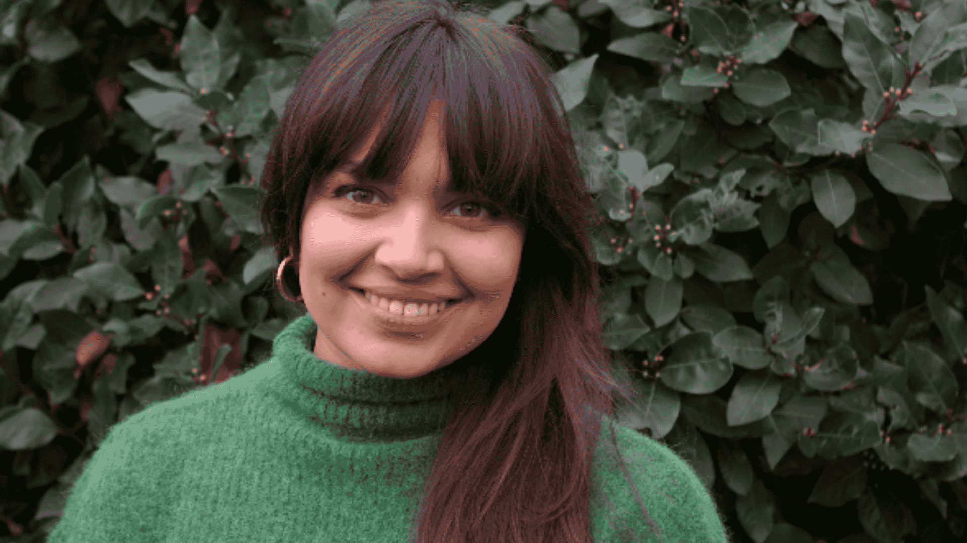 A woman with brown hair styled in a fringe and wearing a green polo neck sweater smiles at the camera. In the background is a bush..
