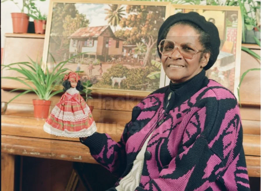 A Black woman smiles at the camera. She is wearing a black hat, pink and black cardigan and glasses. She is holding a child's doll .