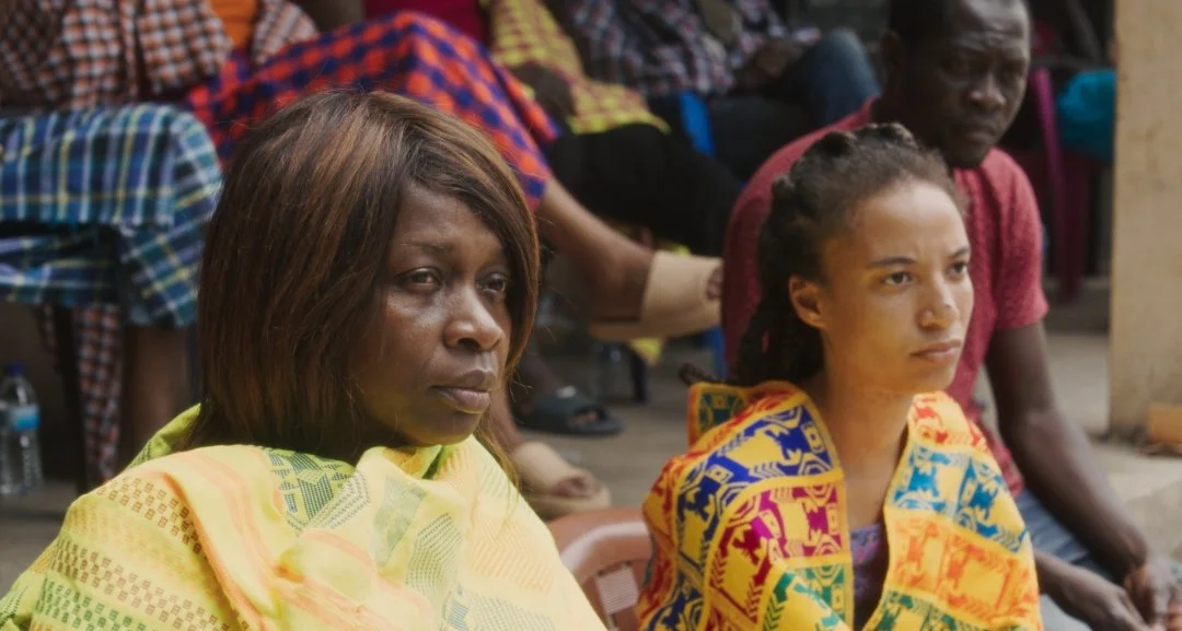 Two black women in traditional african dress sit listening to something out of shot.