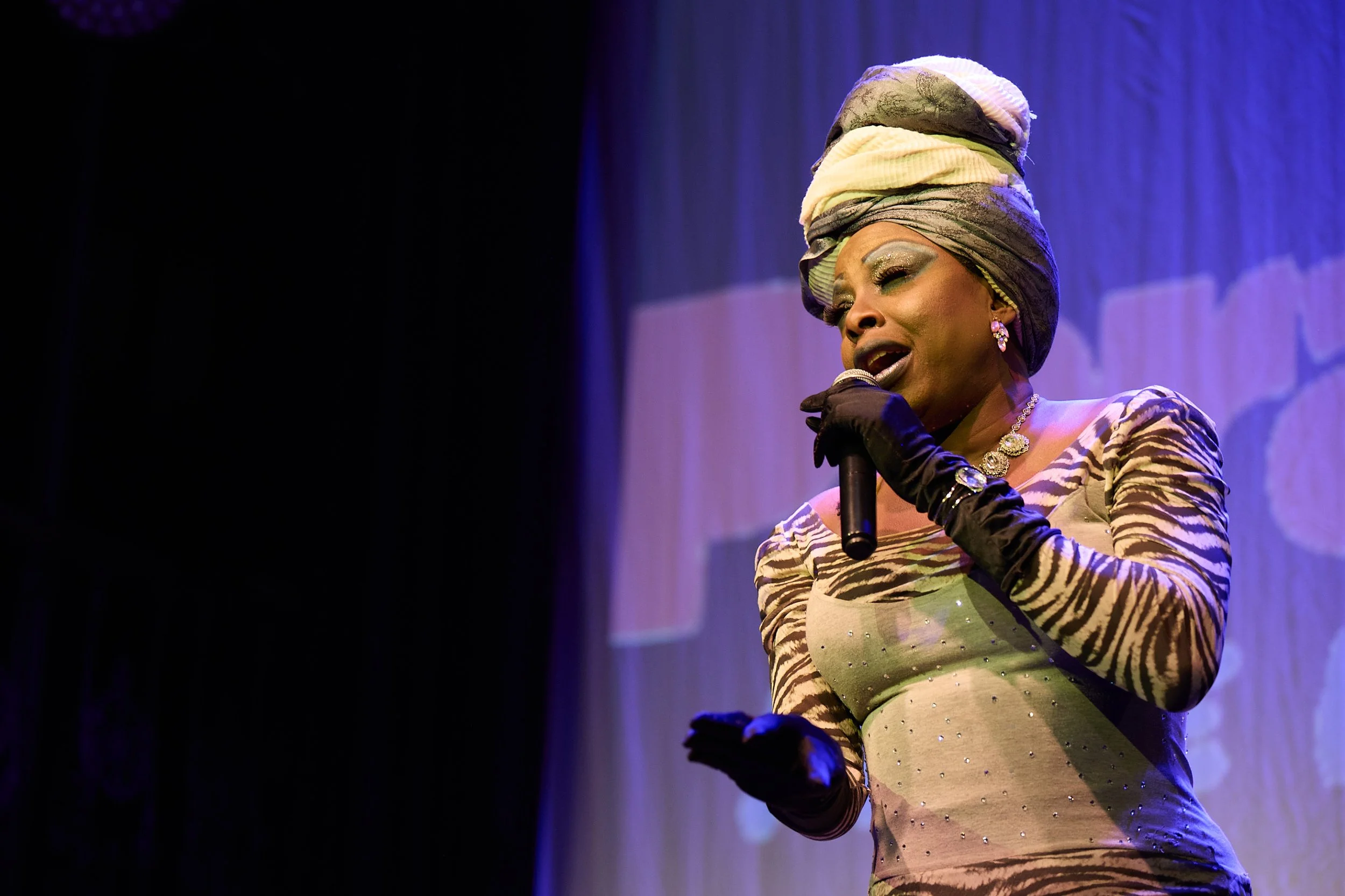 Image of a black drag queen in a head wrap and with a microphone