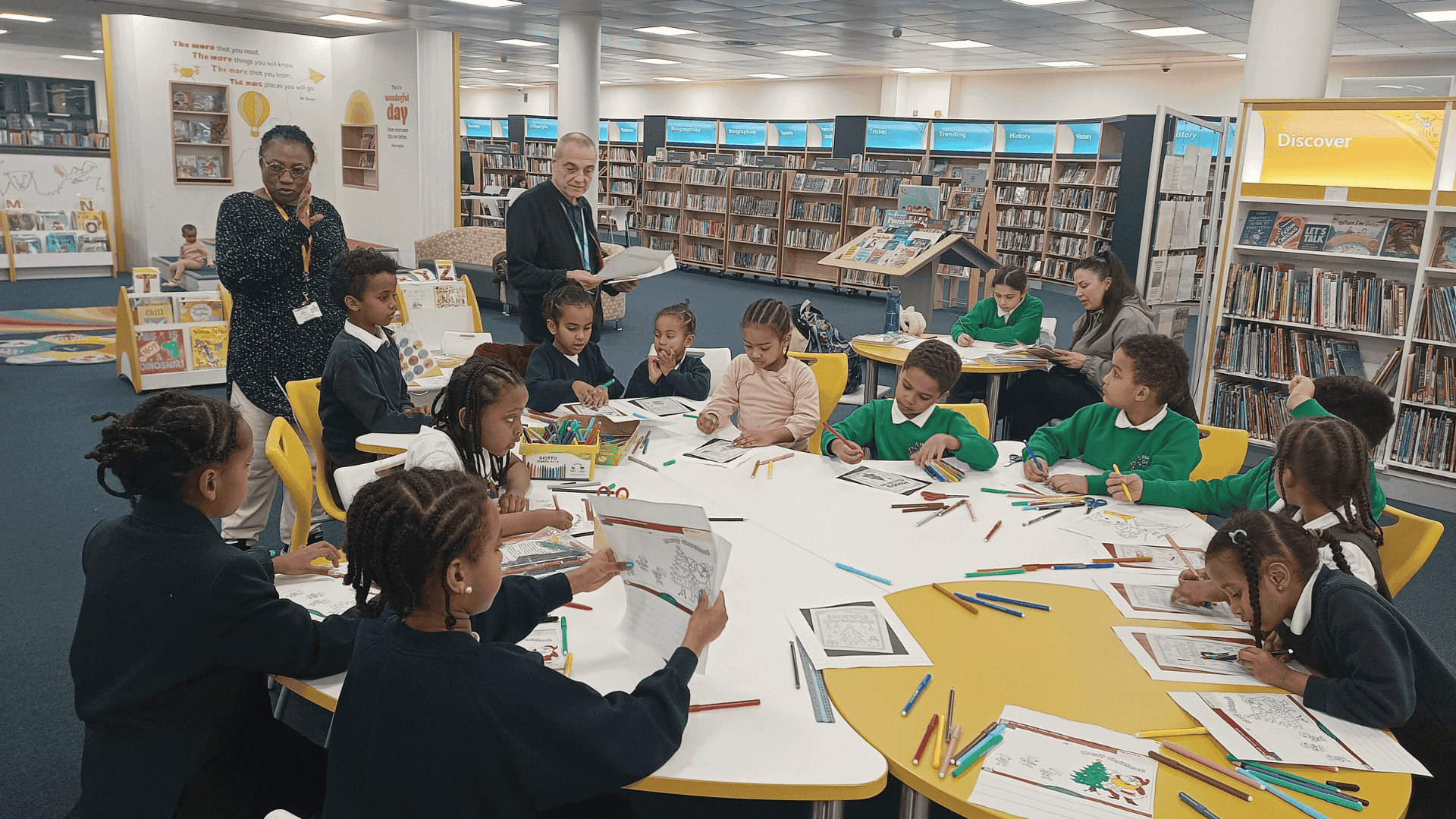 A room - apparently in a library - with primary school age children seated around a table. On the table are several pens and pieces of paper. Two adults stand over them in the background.