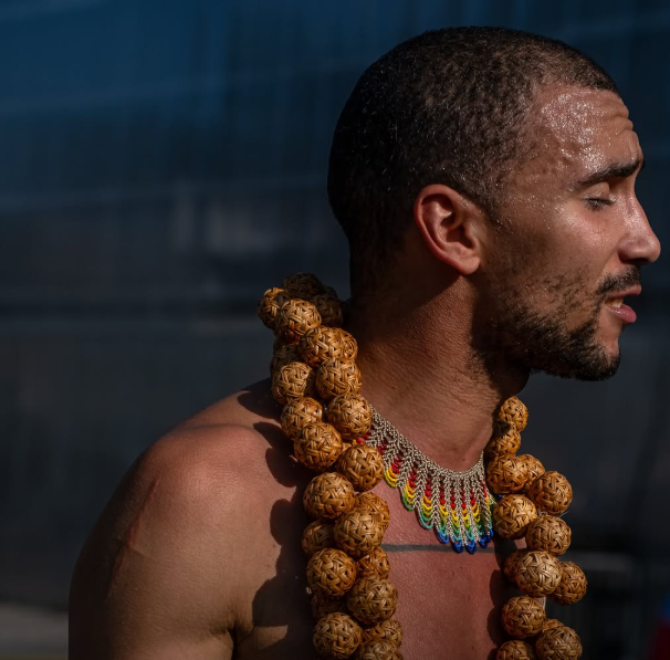 A young POC wears large and colouful jewelry, they perspire like that have been dancing and appear to be topless