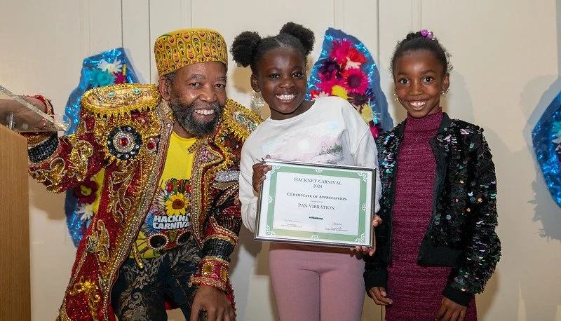 A man in a bejewelled red and gold coat and hat bends down next to two children smiling and holding a certificate