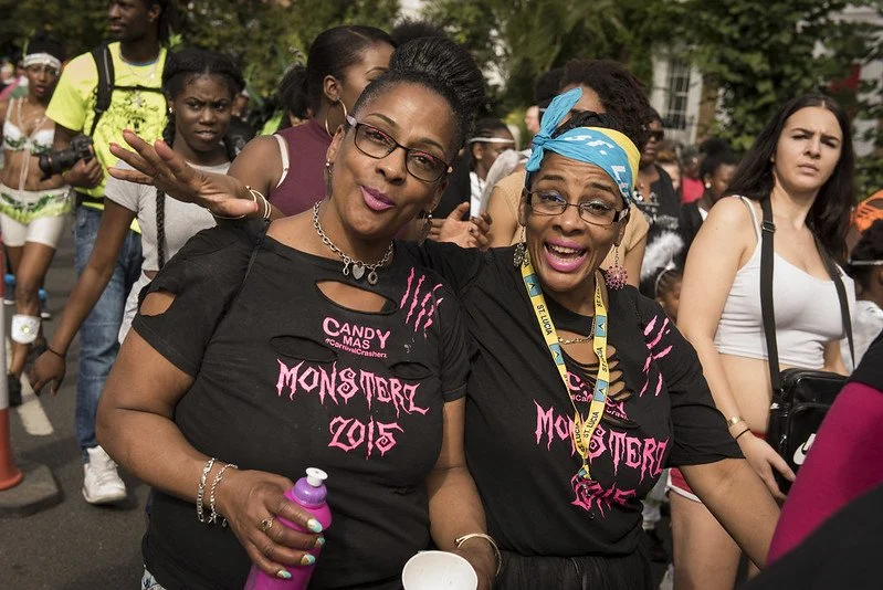 Two women pose for the camera in a carnival parade. The are wearing black t-shirts that have 'MONSTER 2015' written in pink writing.