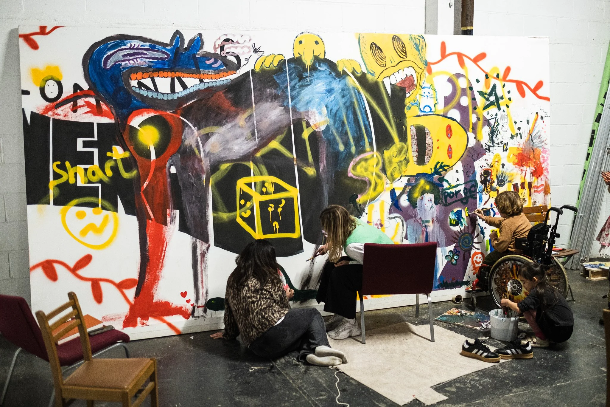 An image of people creating art on a very large white canvas space, four people are shown one is in a wheelchair and one is  a child