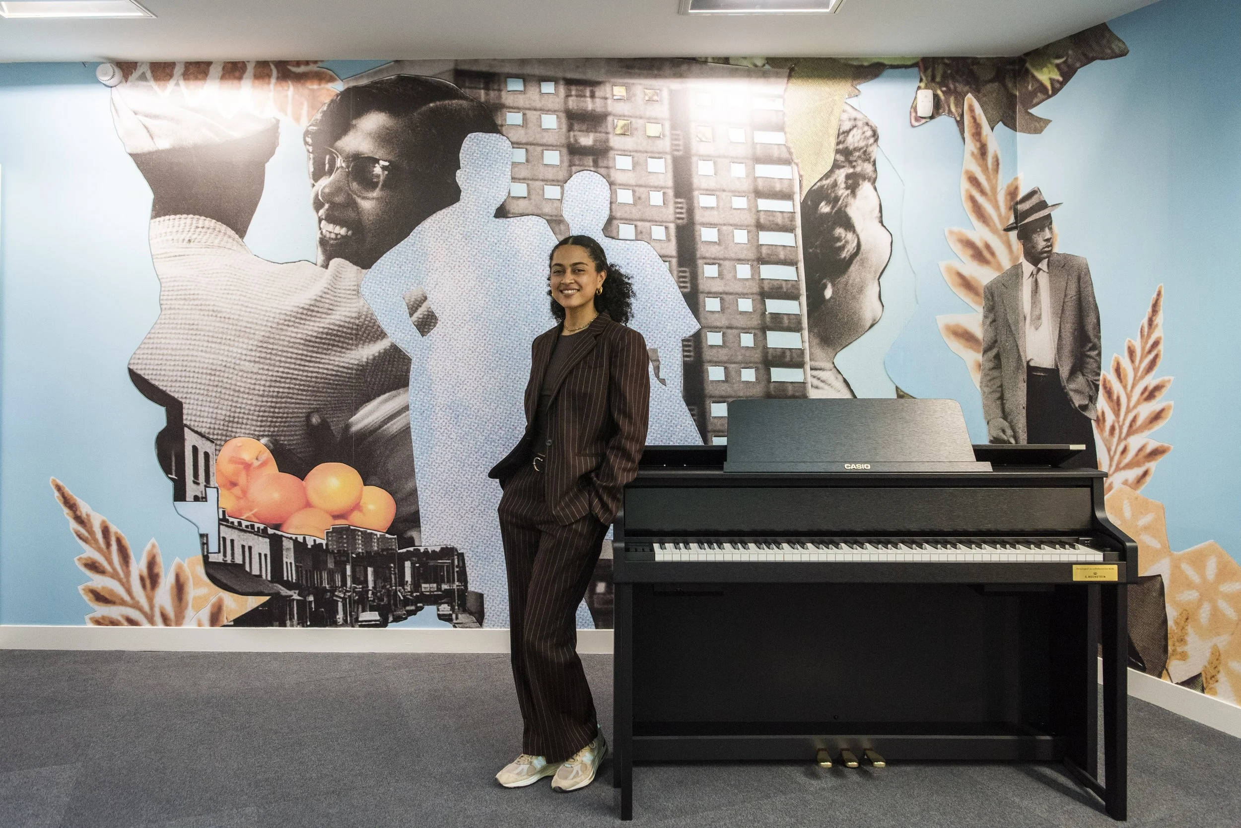 A woman stands infront of a piano and mural. She wears a brown suit and is smiling