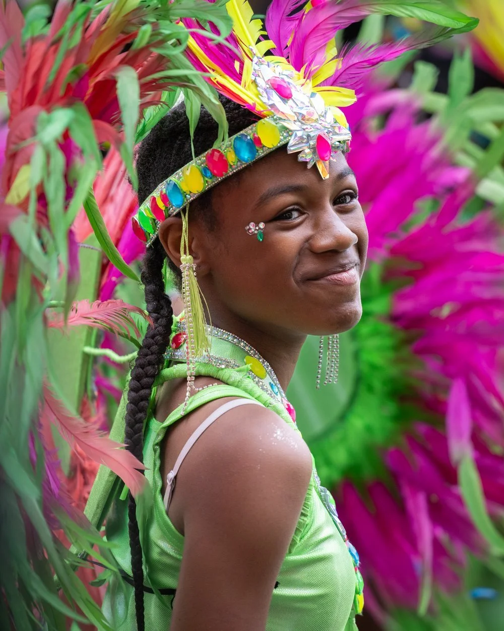 A girl dressed in bejewelled headress and green leotard smiles at the camera. Behind here is a background of green and purple feathers