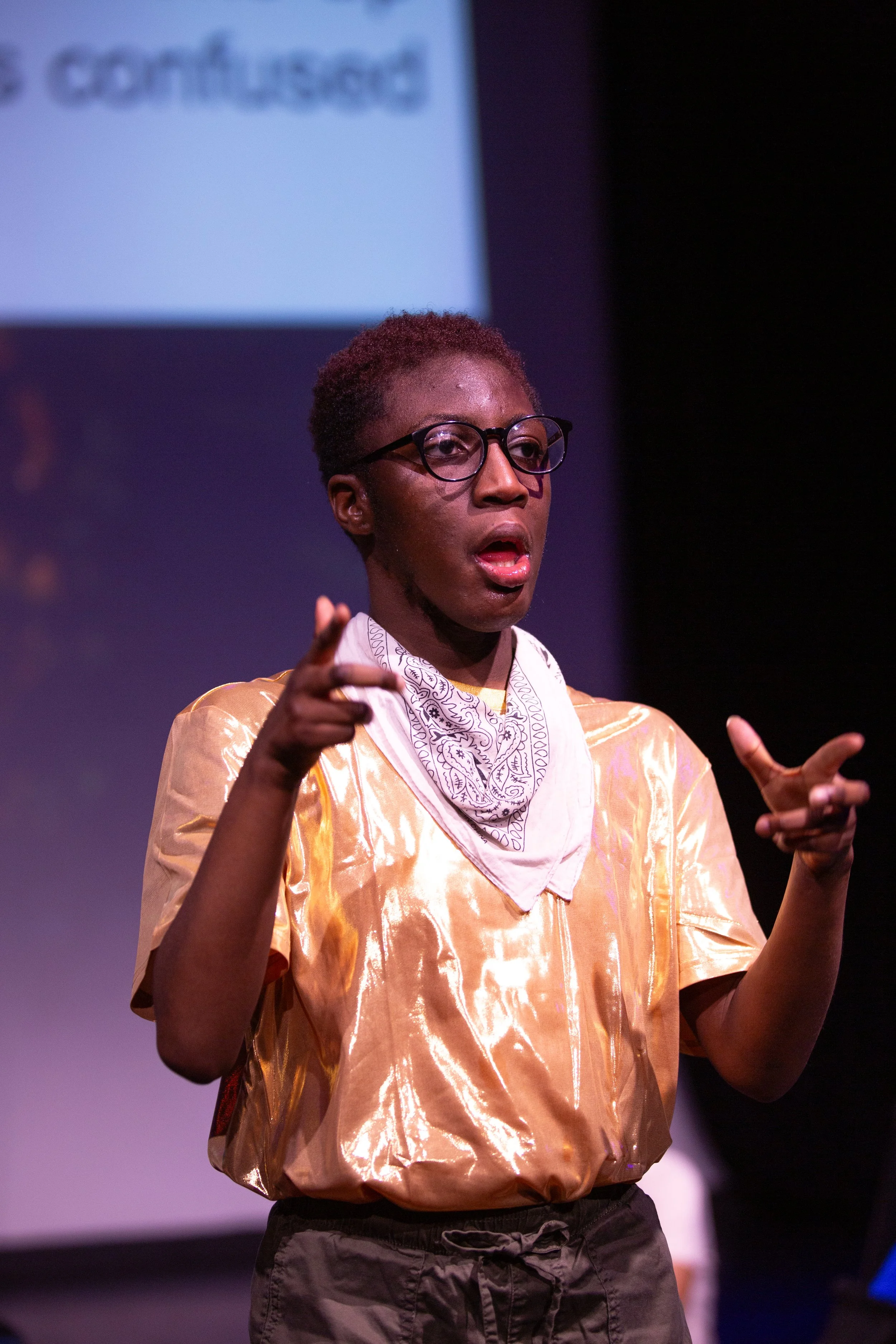 a young black person with round glasses, short hair, a gold shiny top and white bandana around their neck gestures as if acting