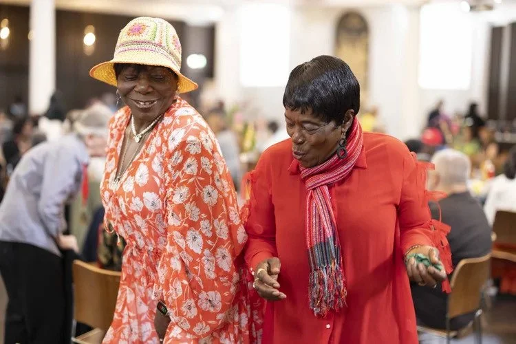 Two Caribbean women dancing. One wears a floral red dress and a summer hat, the other wears a red dress and scarf.