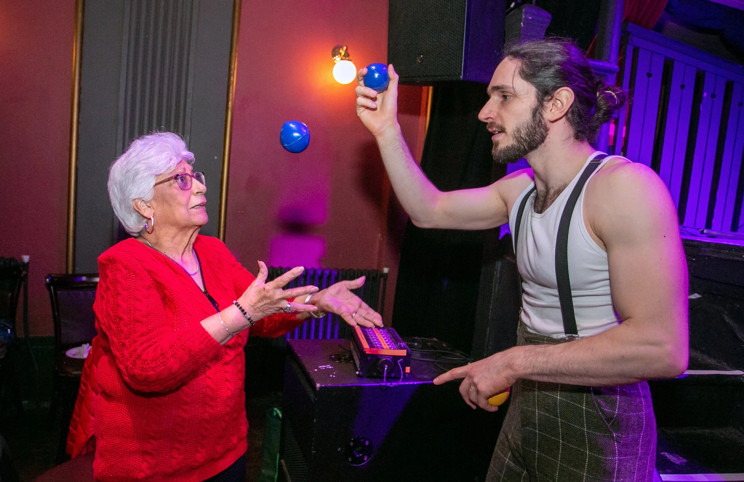 A very happy older woman with grey hair, smiles with her mouth wide open infront or a lourful background, she is wearing a yellow sari. 