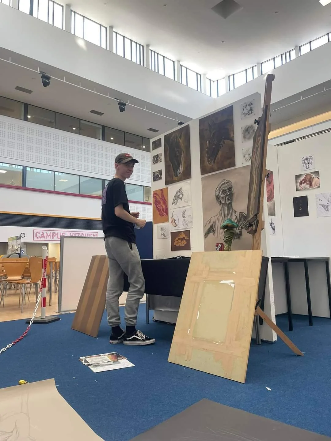 A young person standing next to an art display in an indoor gallery or exhibition space, with various paintings and sketches on the walls behind them.