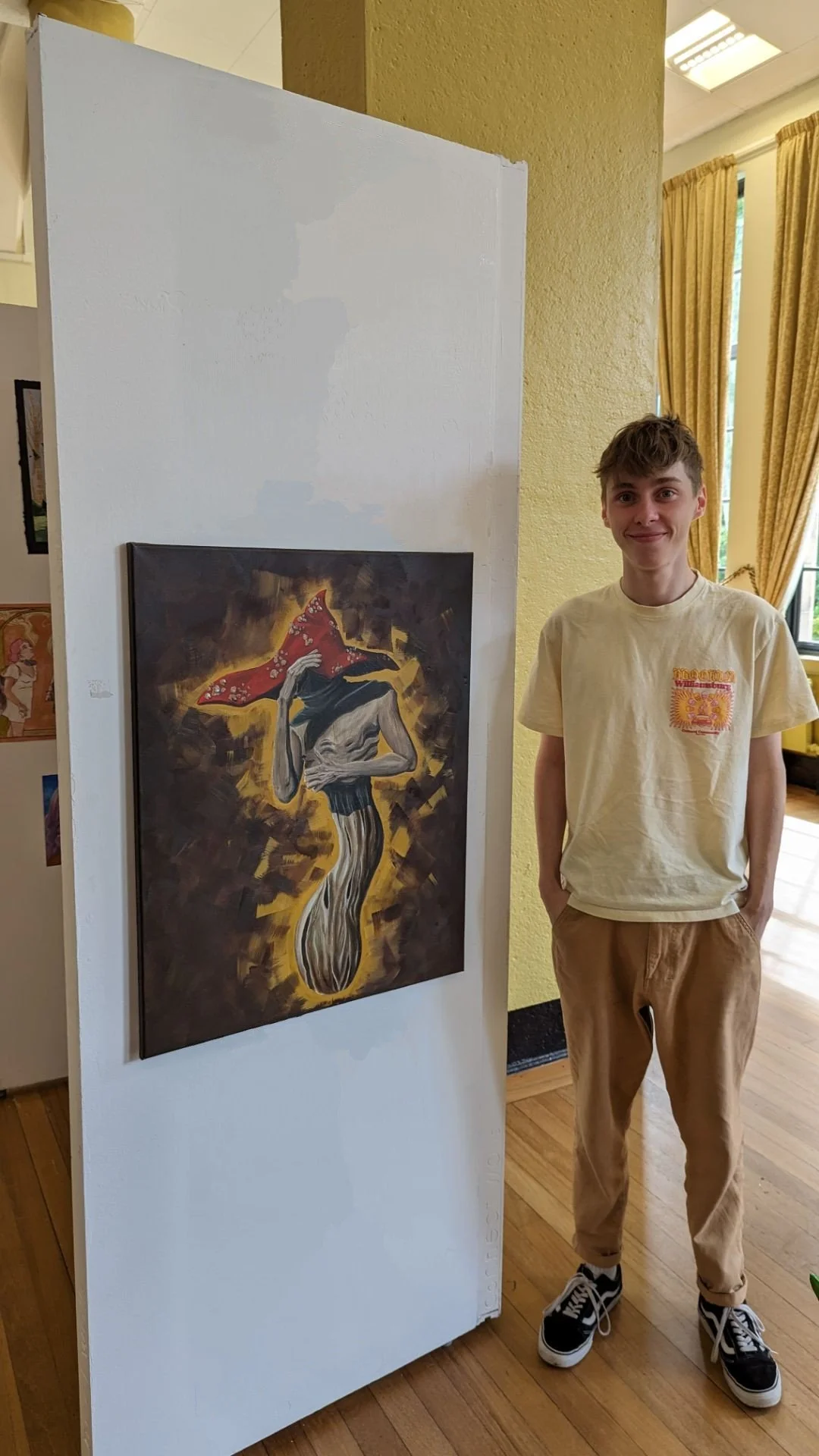 Young man standing next to an art piece in an indoor gallery, smiling, with yellow curtains and a window in the background.