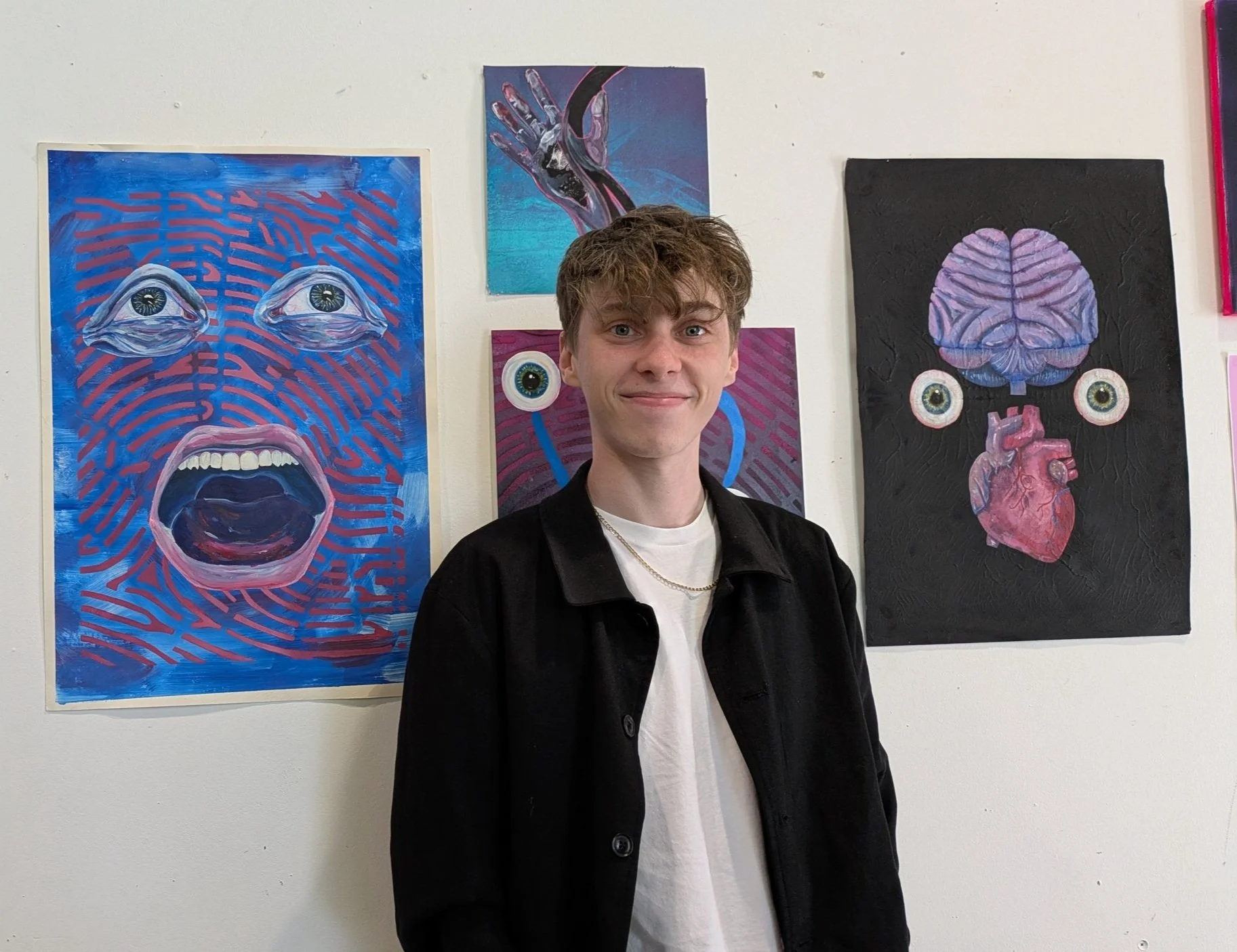 A young man with short, wavy brown hair, wearing a white t-shirt and black jacket, standing in front of an art display on a white wall, featuring paintings with surreal faces and eyes.
