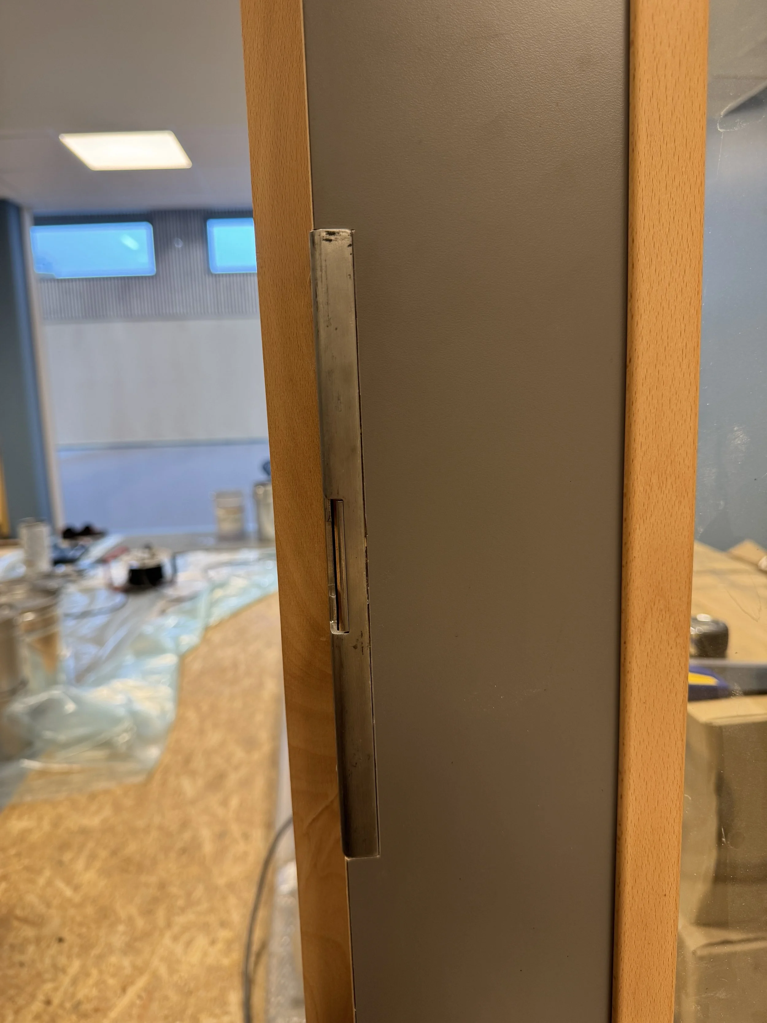 Et bilde av en vegg med en metallbeslag, sannsynligvis en lås eller beslag, og en del av et rom med vinduer og uklar innredning.