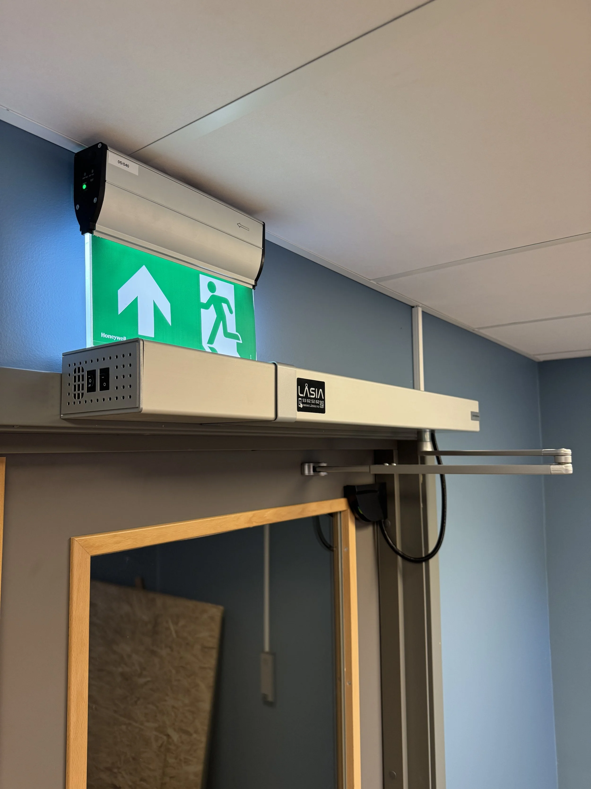 En grønn nødutgangssignale med en oppadgående pil og en illustrasjon av en person som løper ut, plassert over en dør med en automatisk døråpner.