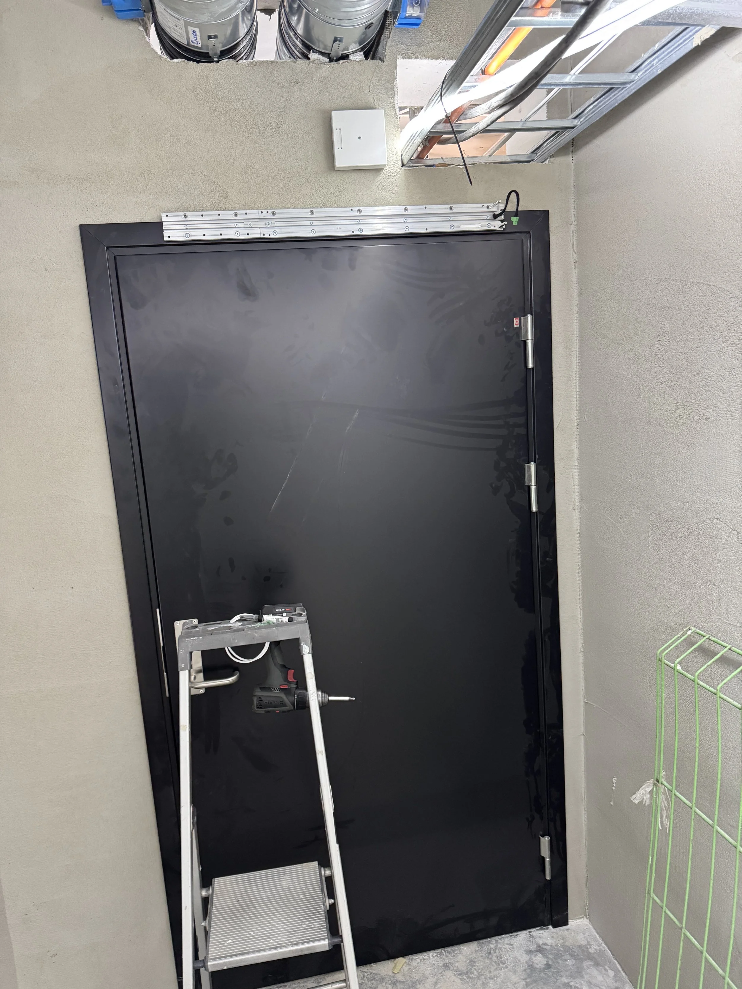 Sort dør installert i en vegg, det er en stige foran døren, og noen elektriske komponenter og rør er synlige rundt døren i et bygge- eller installasjonsområde.