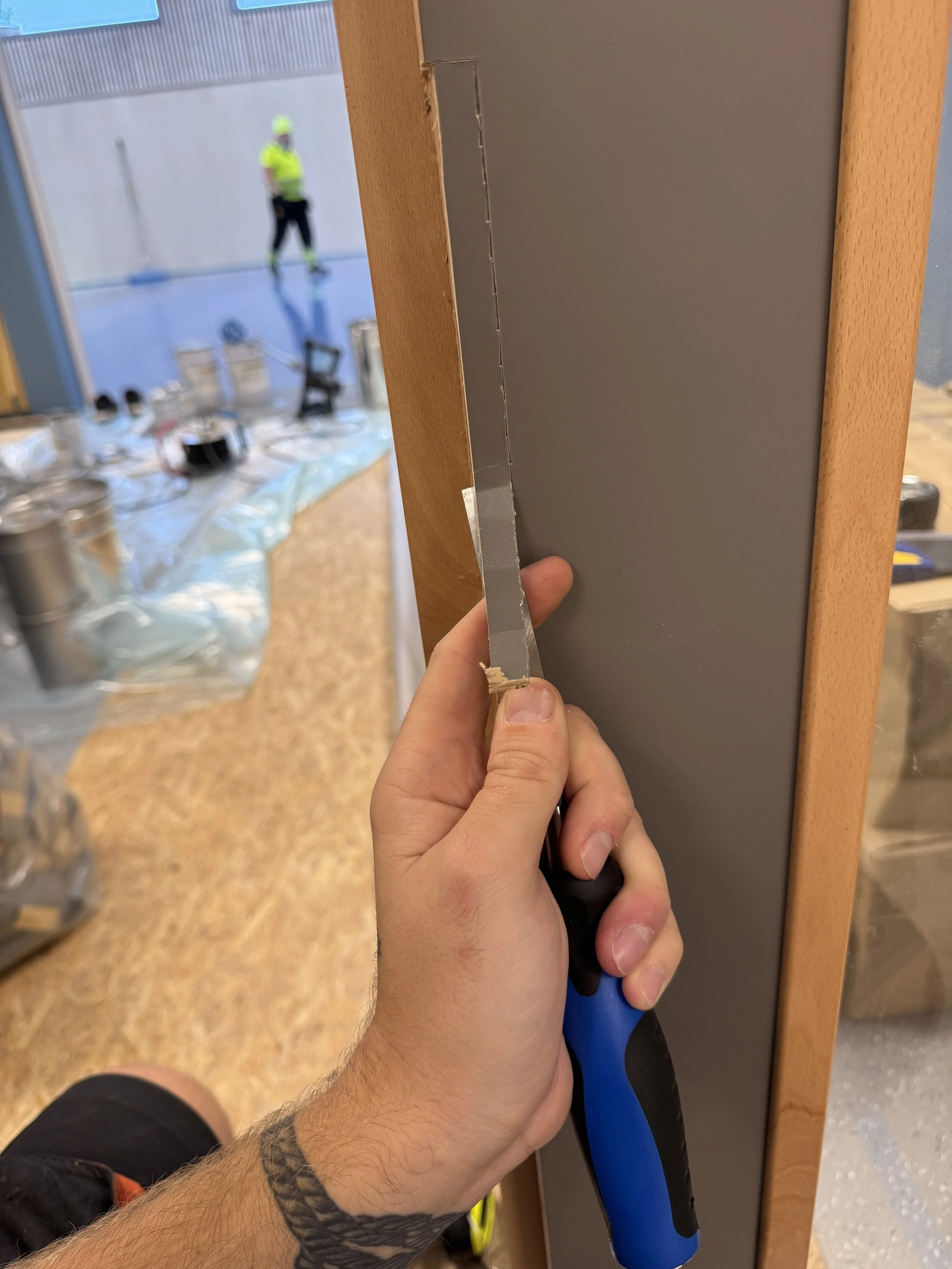 En person holder en håndsag og skjærer i en veggkarm av tre, mens en byggeplass eller verksted er i bakgrunnen.