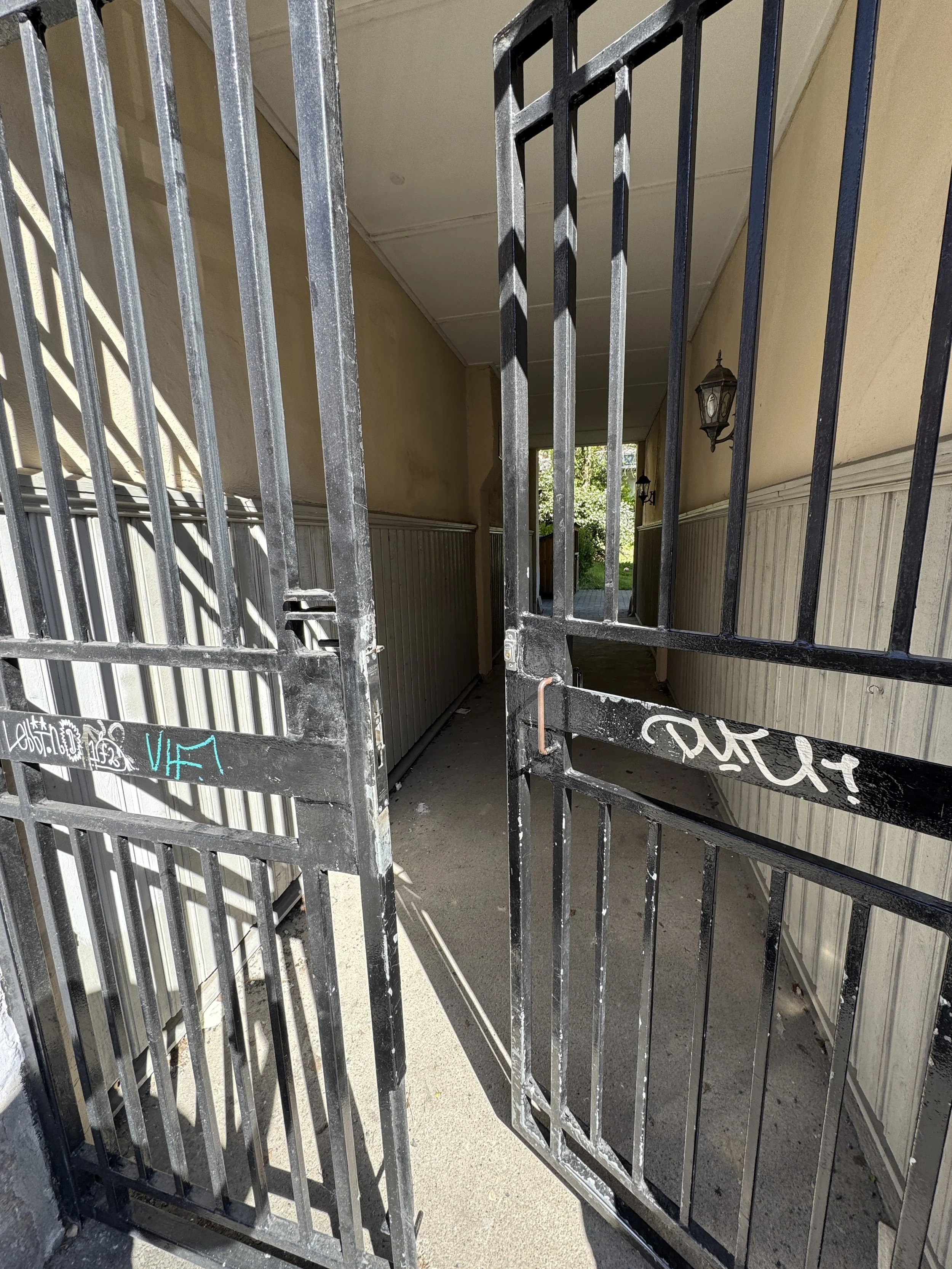 Åpen svart metallgitterport som leder inn til en inngang under tak, med en vegg med lysere panel og utendørs trær i bakgrunnen.