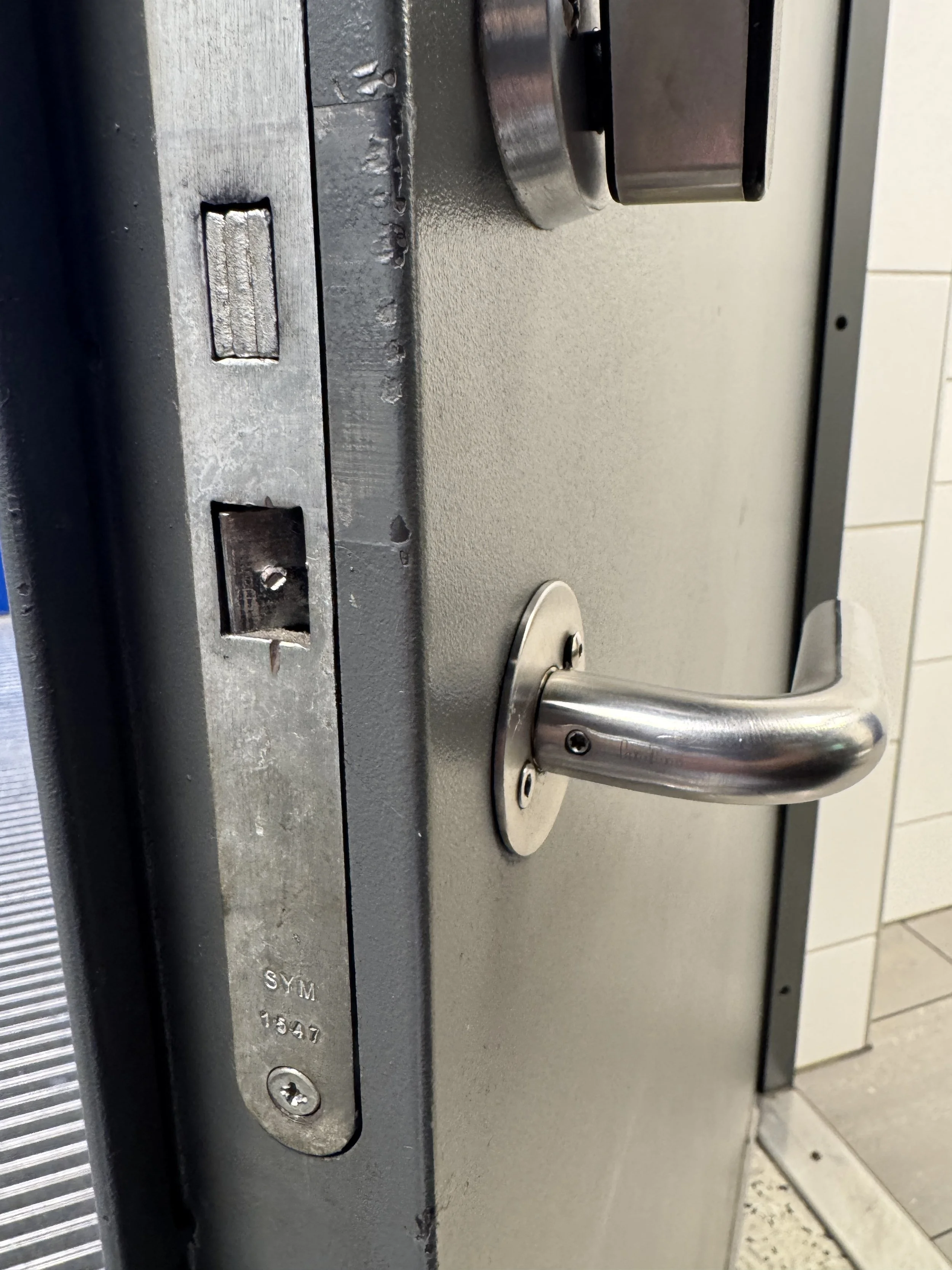 En metall-legearm, som er en del av en låsemekanisme på en dør, sett fra siden. Døren er grå, og det finnes en personbeskyttet mekanisme for å låse opp eller låse døren.