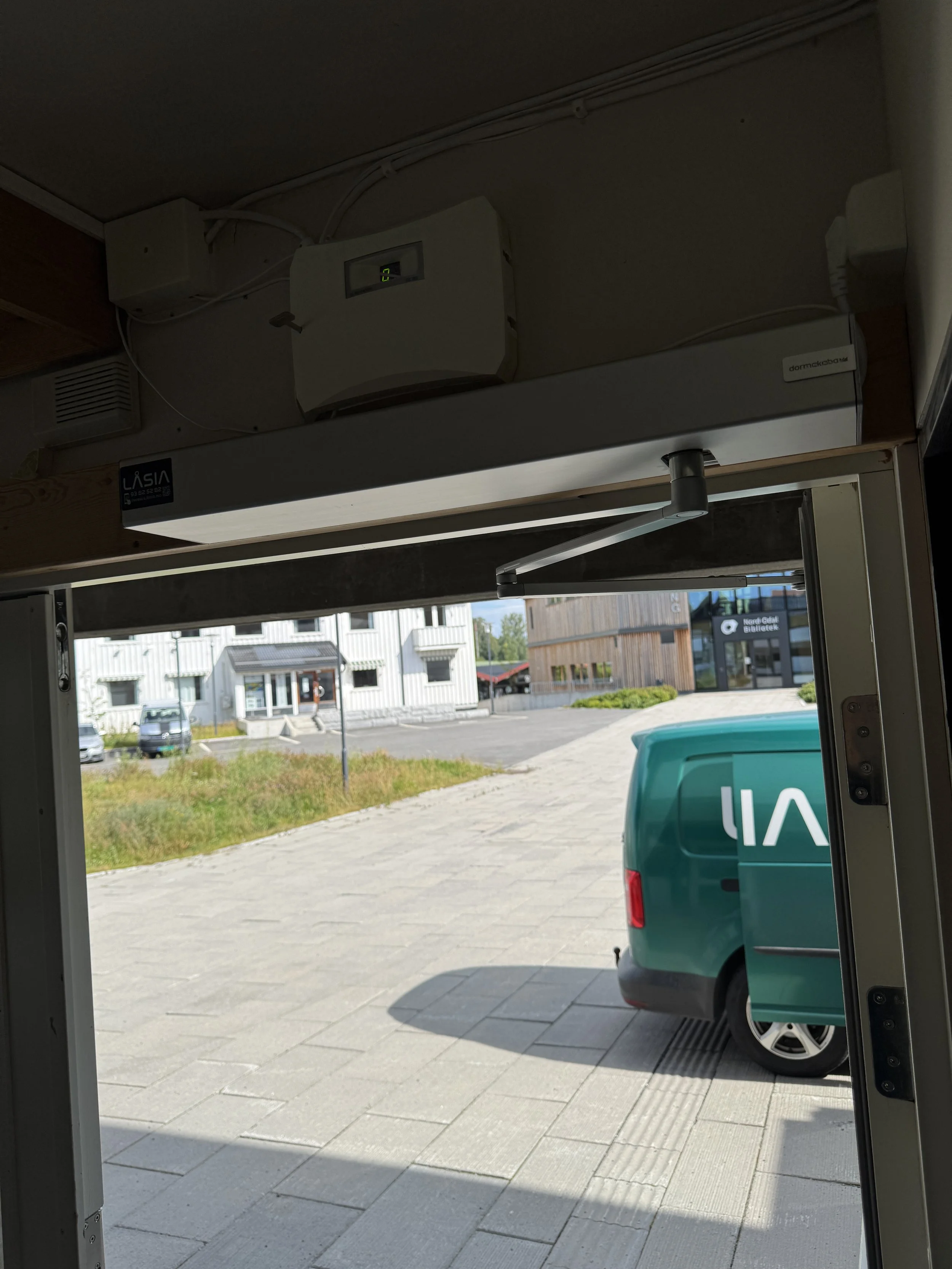 Utsikt ut gjennom en dør mot en parkeringsplass med en grønn varebil og noen bygninger i bakgrunnen.