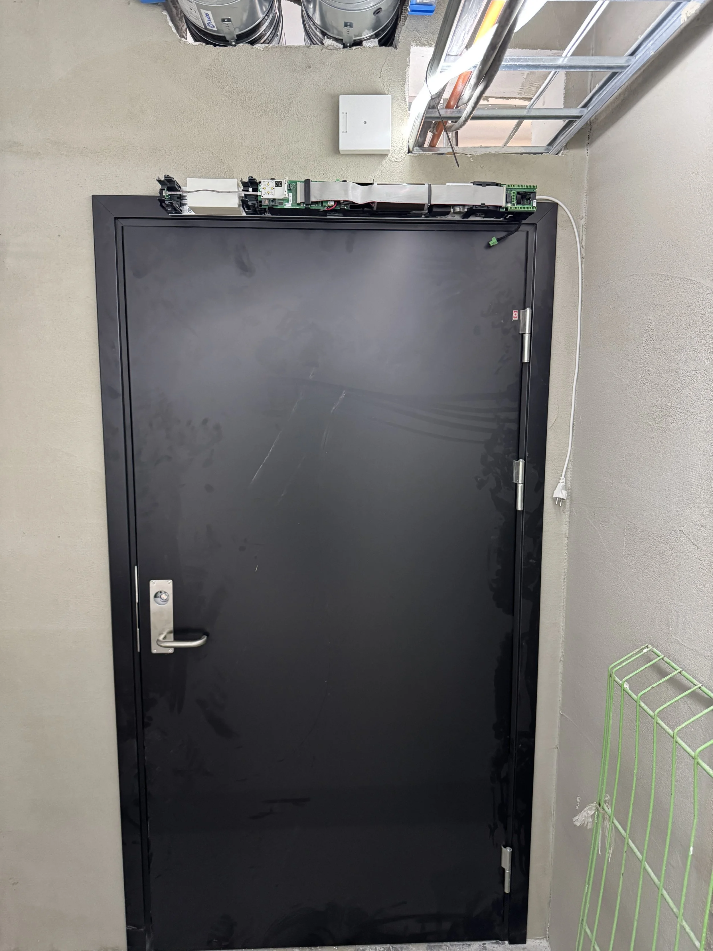 En svart sikkerhetsdør med synlig mekanikk og elektriske komponenter over øvre del, plassert i et rom med beige vegger. Dette ser ut til å være en del av en teknisk installasjon eller kontrollrom.