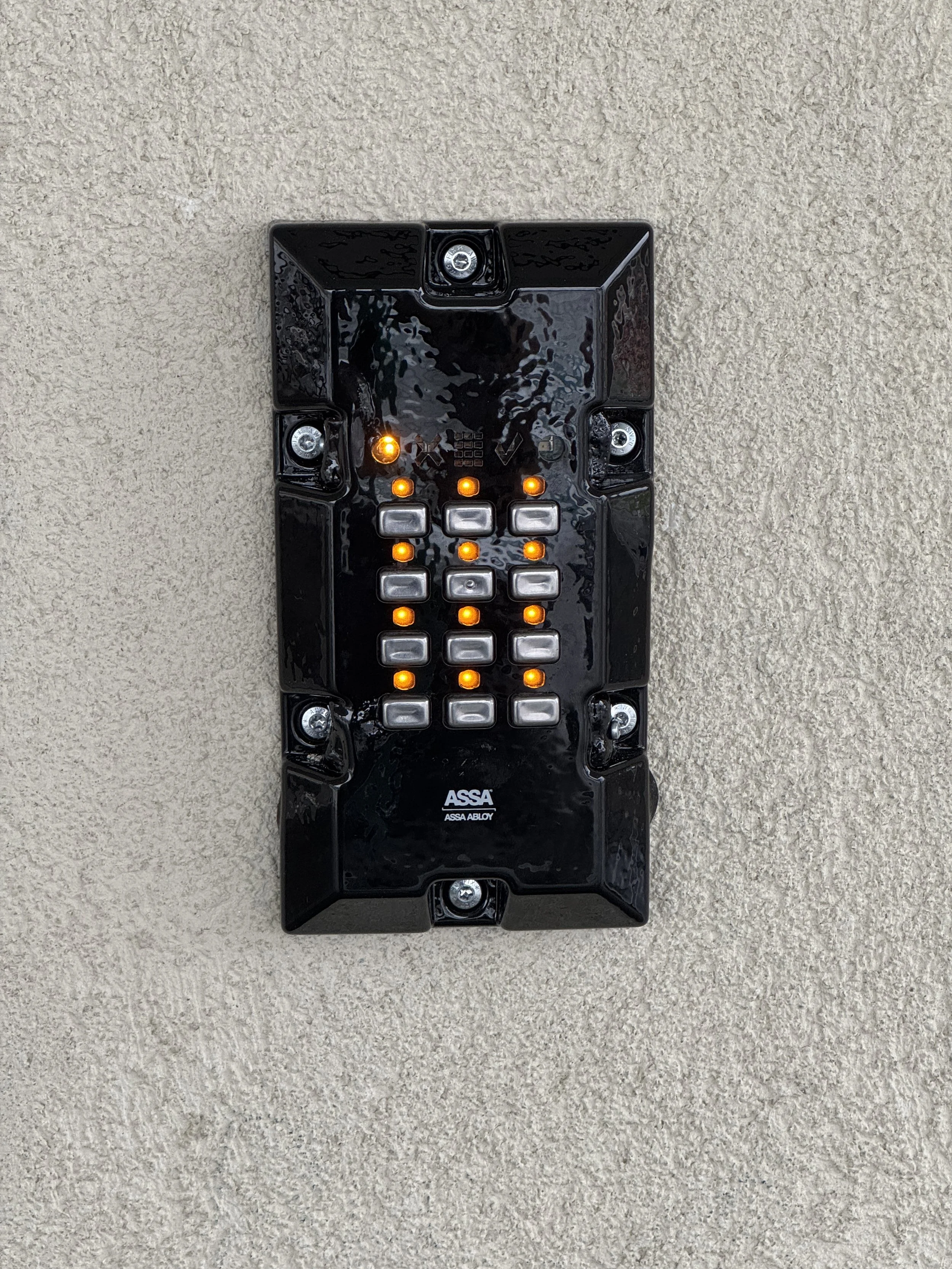 Sort nødtelefon- eller alarmboks med lyse oransje lys, montert på en beige vegg.