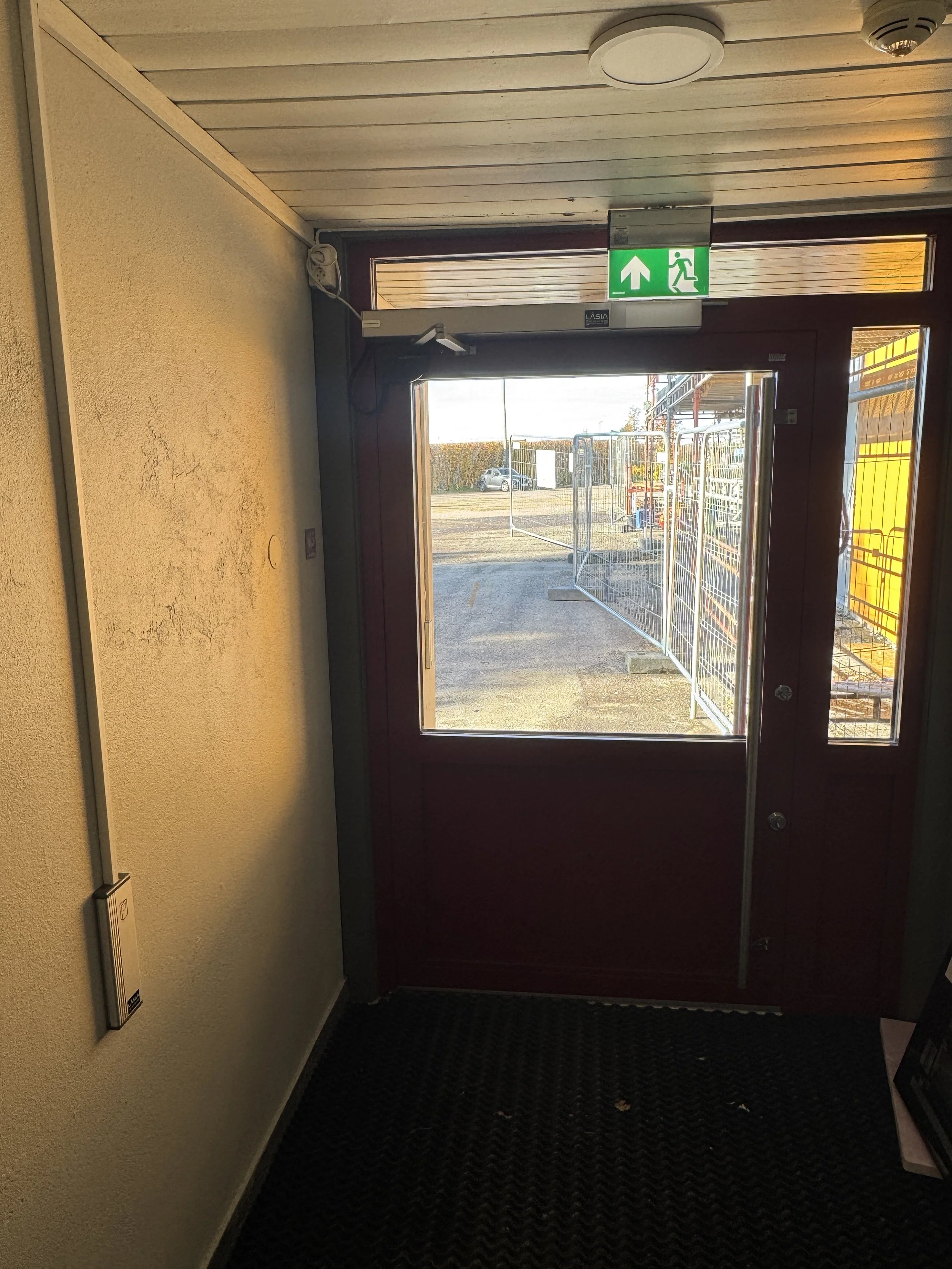 Innendørs bilde av en inngangsdør med en grønn nødutgangslampe over, som peker opp og mot utgangen, med utsikt utendørs til en parkeringsplass og gjerder.