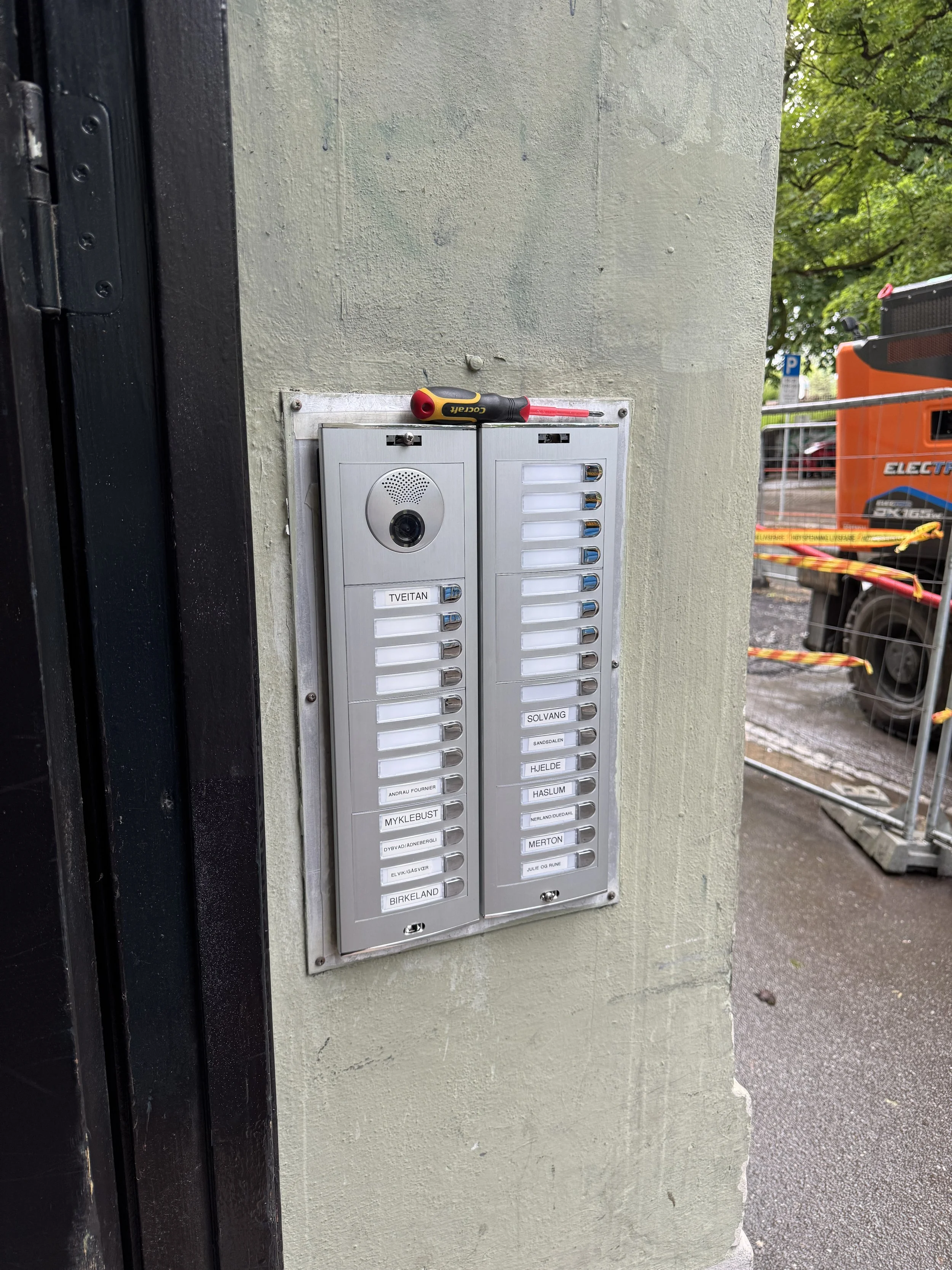 Relé- og porttelefonpanel med flere navnelapper og en skrutrekker liggende på toppen, montert på en vegg utendørs.