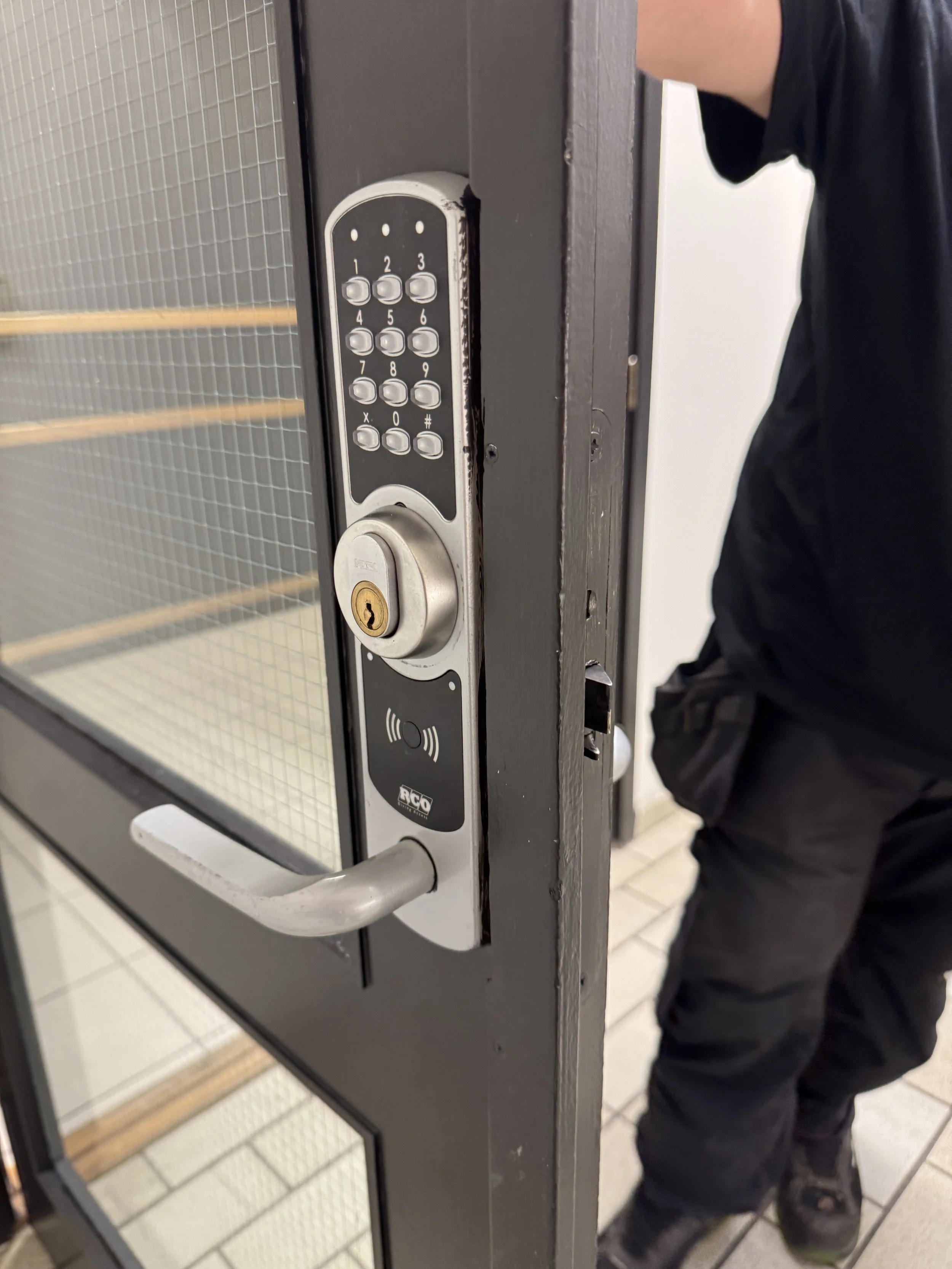 En svart dør med elektronisk lås og tastatur, og en person i svart klær står til høyre.
