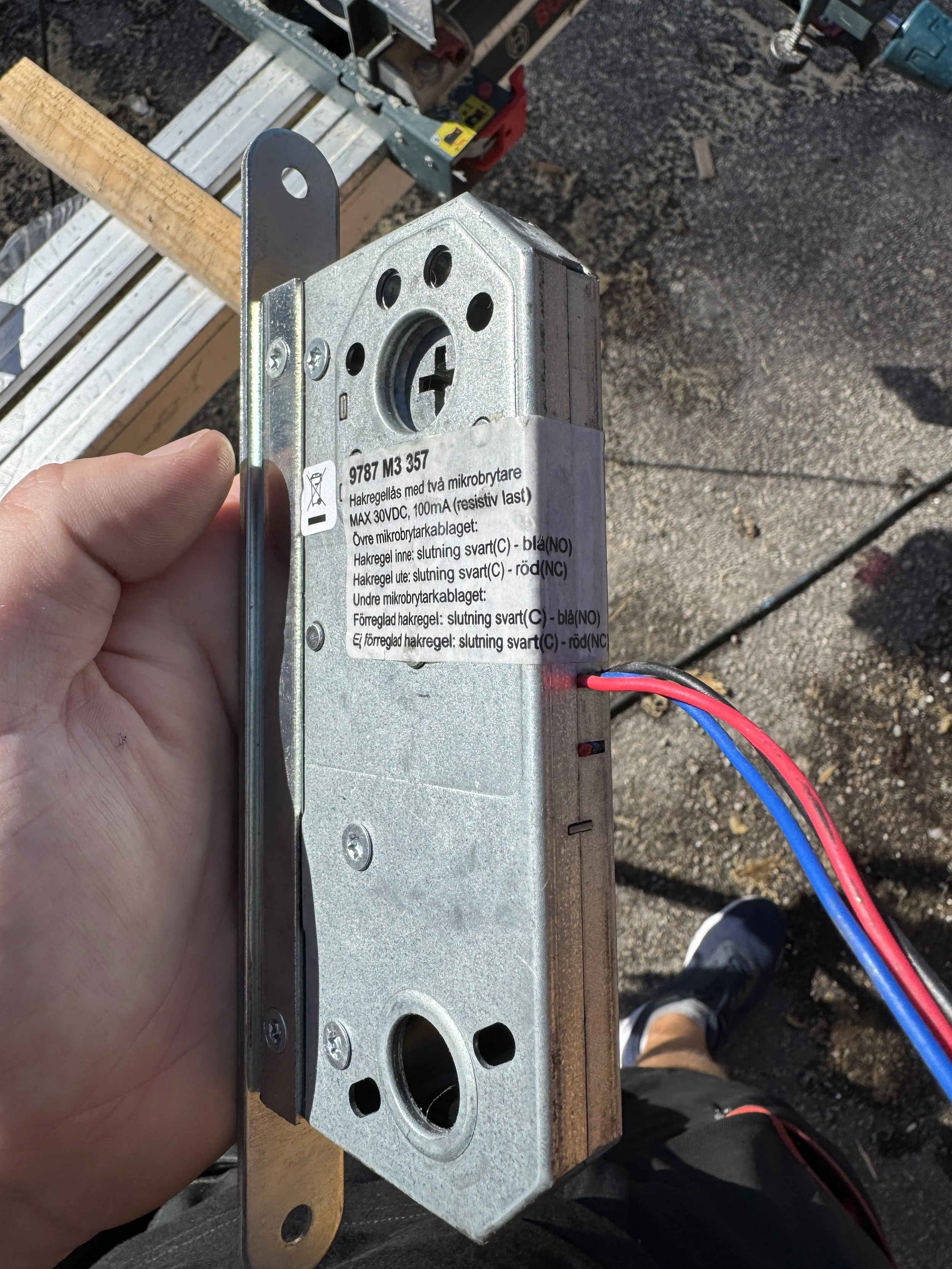 Bildet viser en person som holder en elektrisk komponent med røde og blå ledninger, sannsynligvis en lås eller elektromagnetisk mekanisme, med byggeutstyr og en arbeidsplass i bakgrunnen.