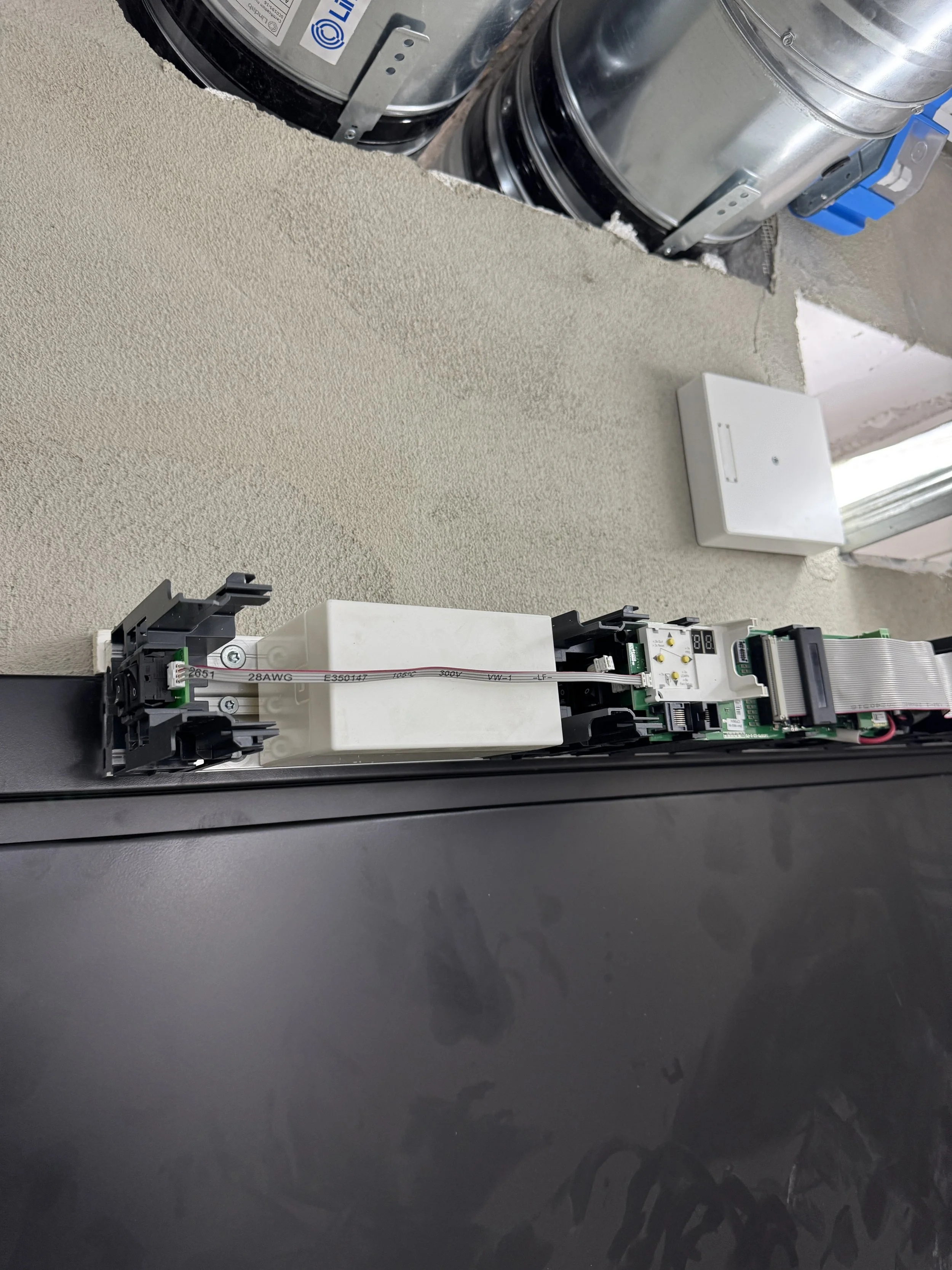 Elektrisk komponent installert på en mørk overflate, med synlige koblingskabler og en LCD-skjerm, plassert på en bygningsvegg.